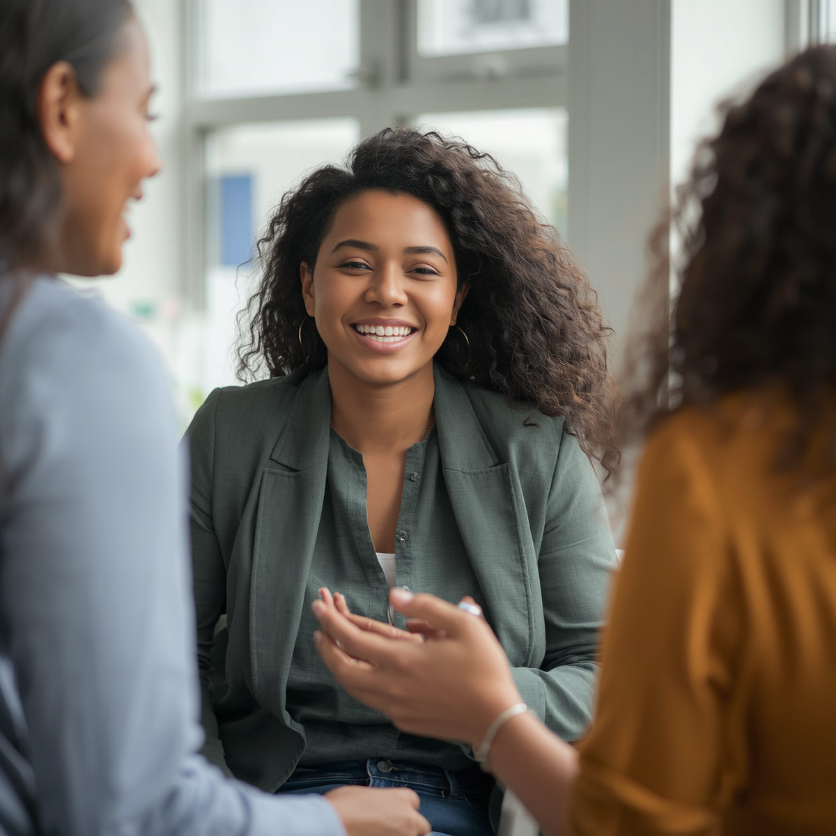 A medium close-up shot of a young Black woman with long, curly dark hair sitting in a brightly lit meeting space. She is wearing a dark olive green blazer over a matching button-down shirt and is smiling warmly and directly at the camera. In the foreground, the soft-focus silhouettes of two other women are visible—one in a light blue shirt on the left and another in a mustard yellow top on the right—gesturing with their hands as if engaged in an encouraging group conversation or networking session.