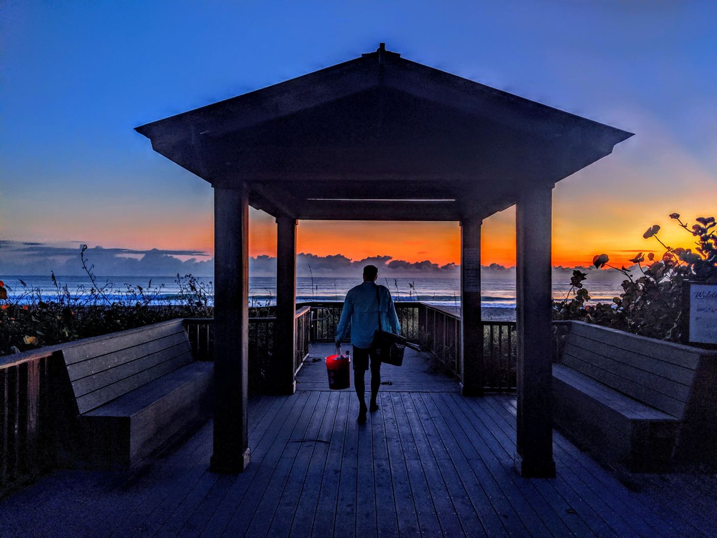 Beachgoer walking through a wooden beach pavilion toward the ocean at daybreak