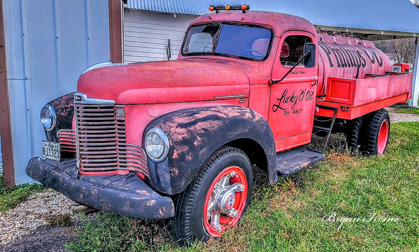 Vintage red tanker truck parked on grass beside a metal barn