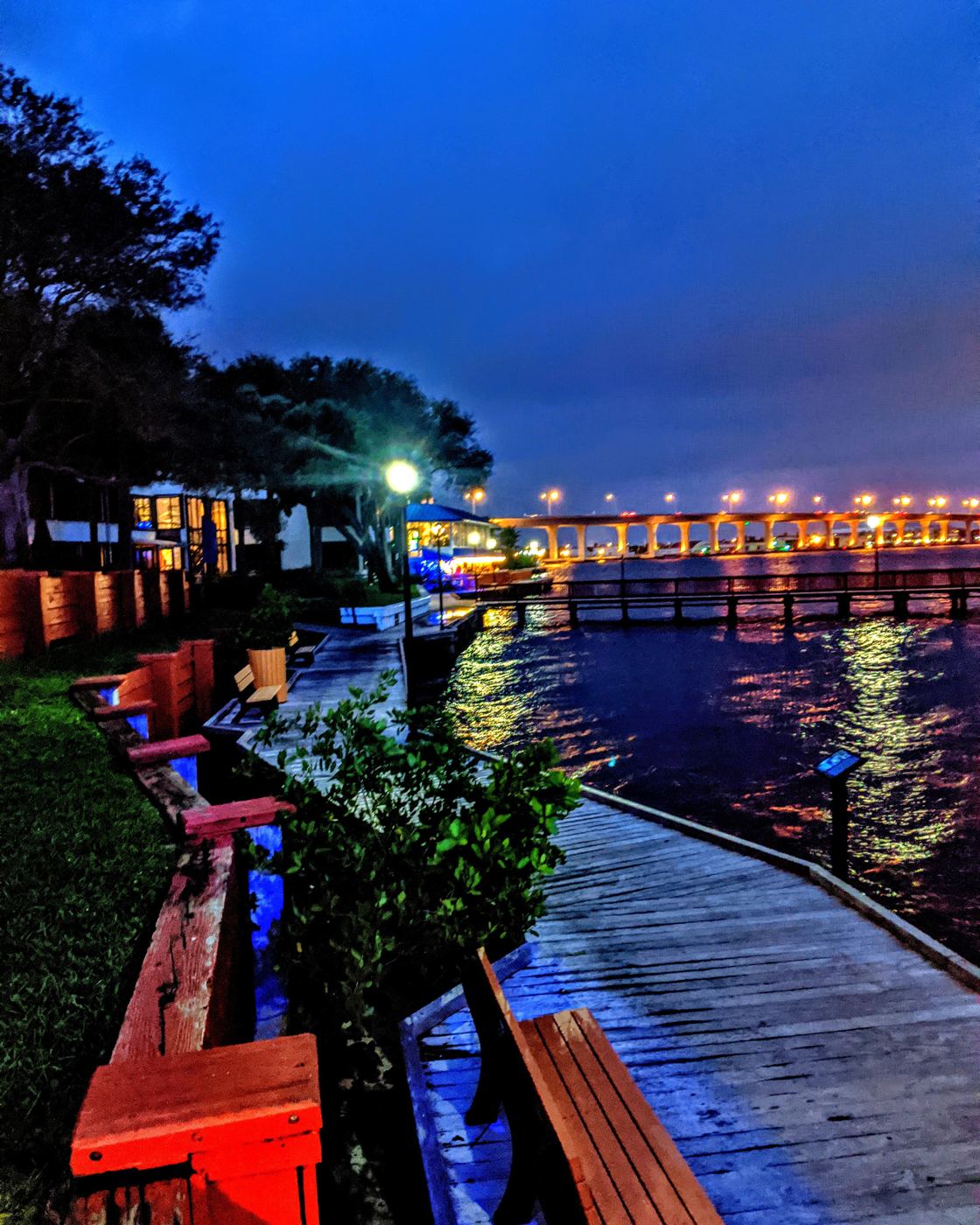 Riverfront boardwalk with red benches and a lit bridge over the water at night