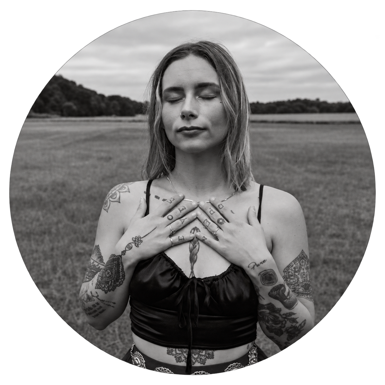 Samantha G in black and white, a blonde woman standing in a field with her eyes closed and her tattooed arms crossed over her chest