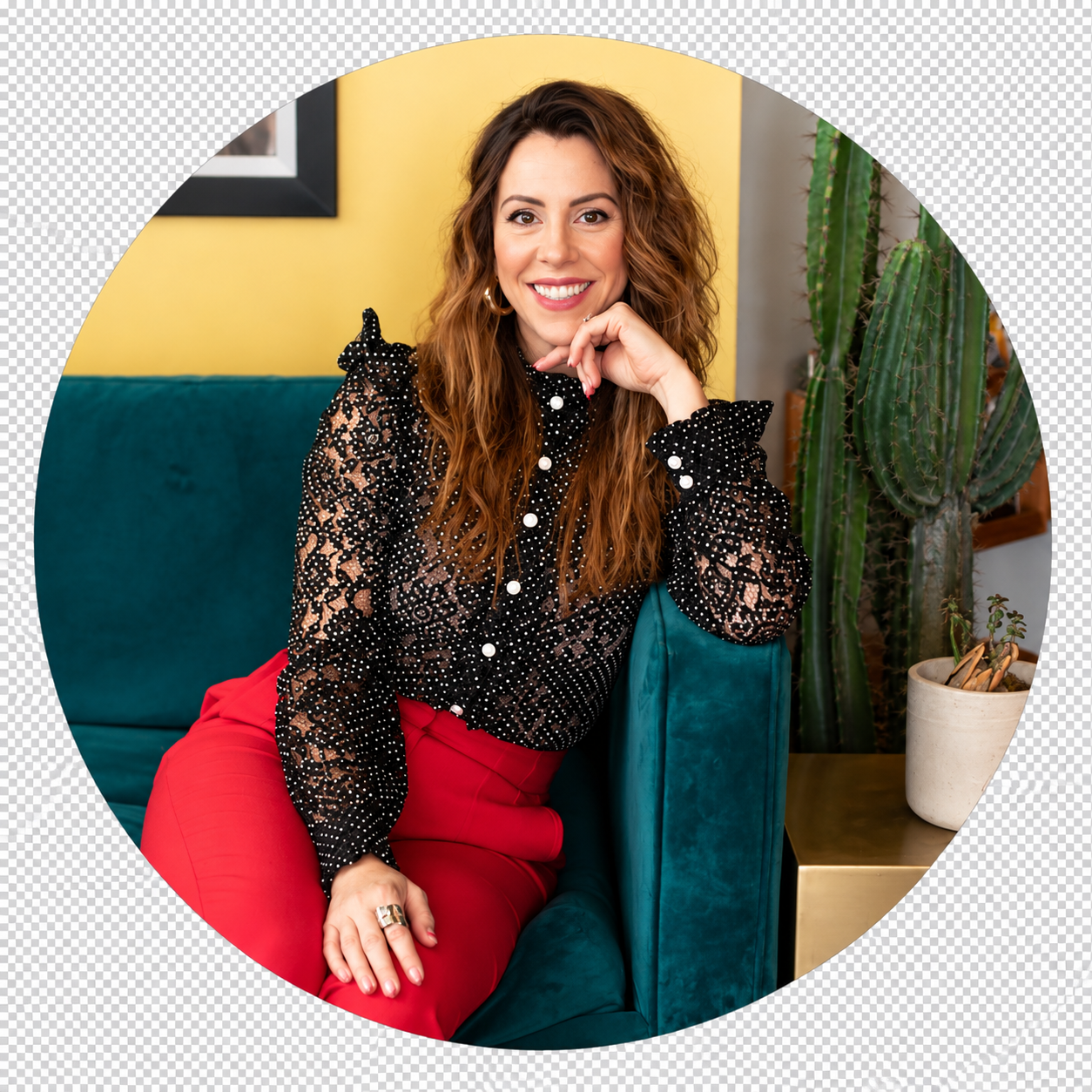 Alisha J, a woman with auburn curls in a black polka-dot lace blouse and red trousers, seated on a teal sofa against a yellow wall