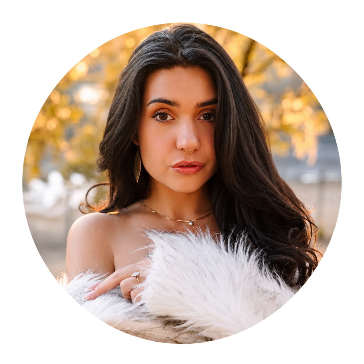 TT, a dark-haired woman with a nose ring and a gold leaf earring, wrapped in white faux fur against golden autumn light
