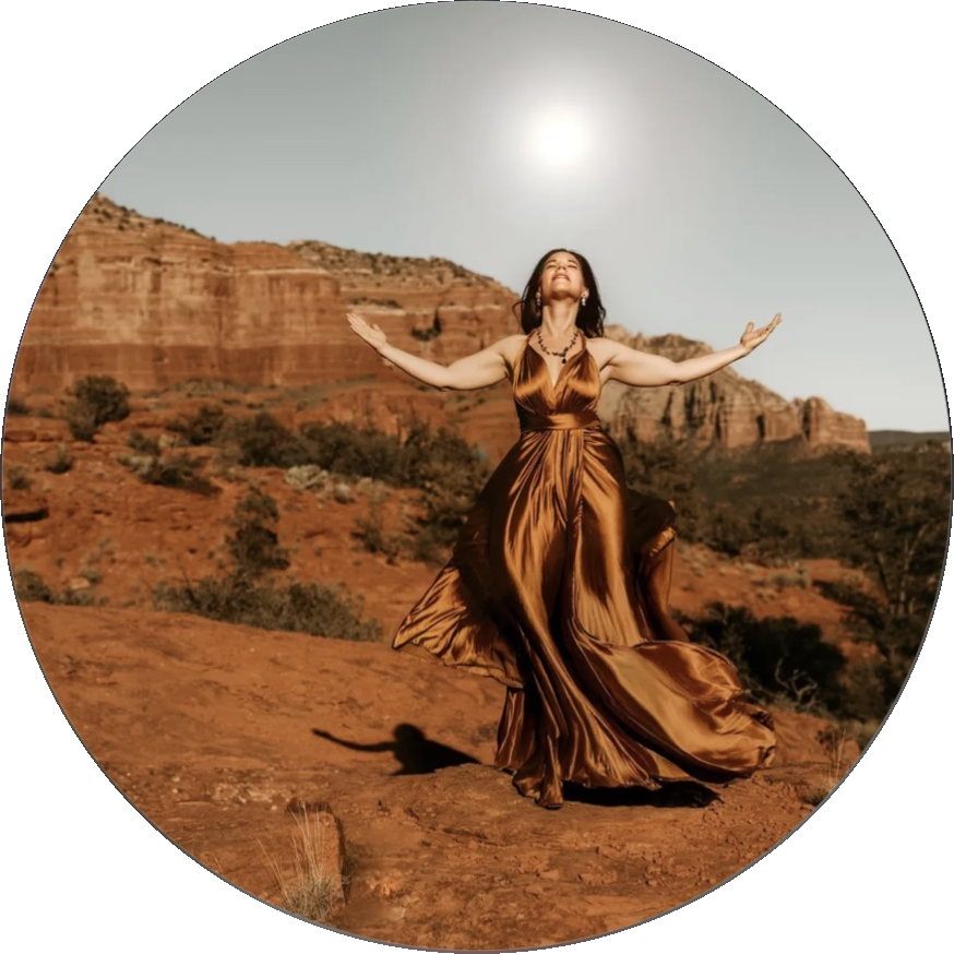 A woman in a bronze satin gown standing with her arms flung wide and her head tipped back in a red rock canyon under bright sun