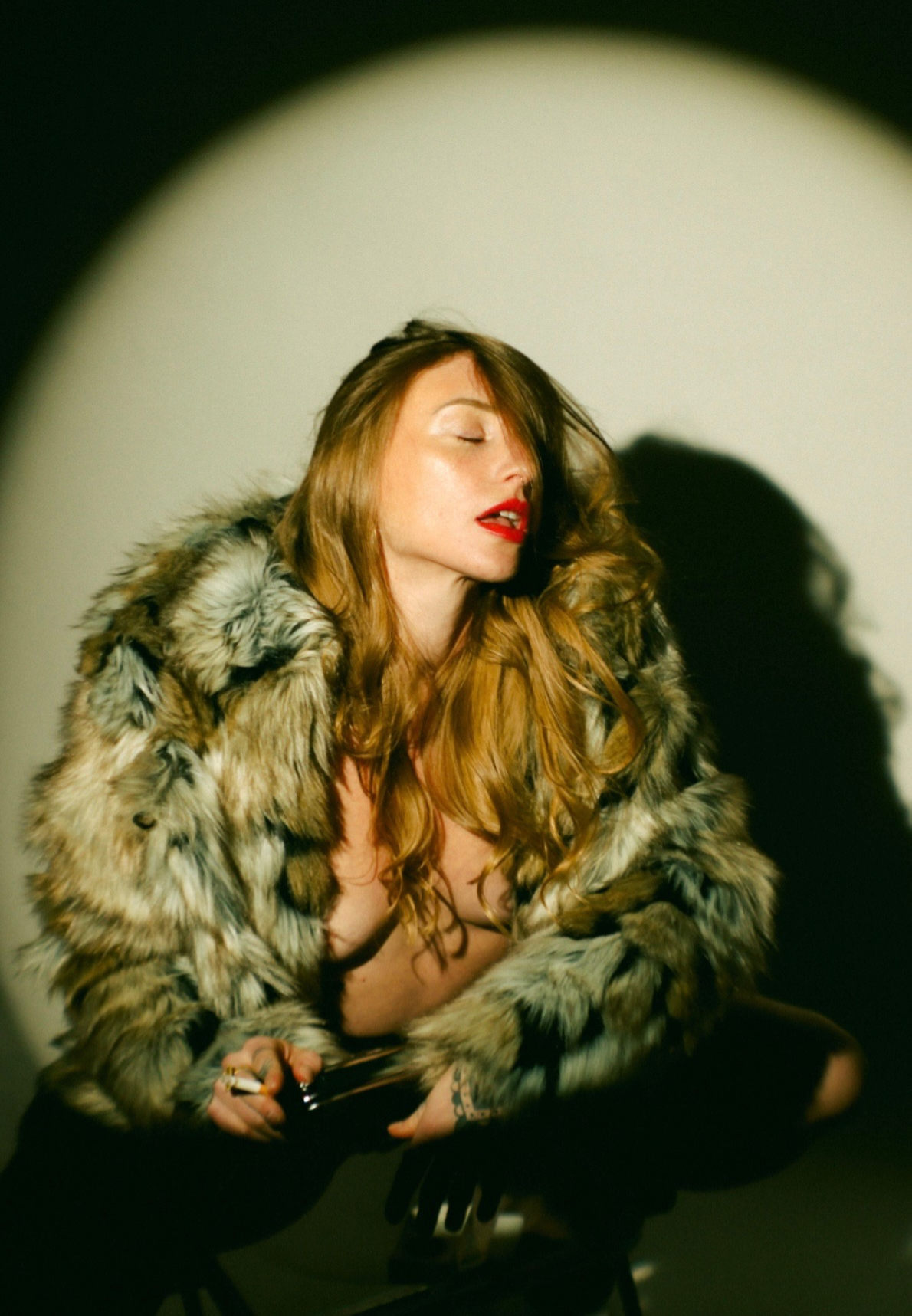 A woman in an open mottled fur coat over her bare chest, red lipstick, her head tipped back and eyes closed, crouched on a metal stool with a cigarette between her fingers as a hard spotlight throws an arched shadow on the cream wall behind her