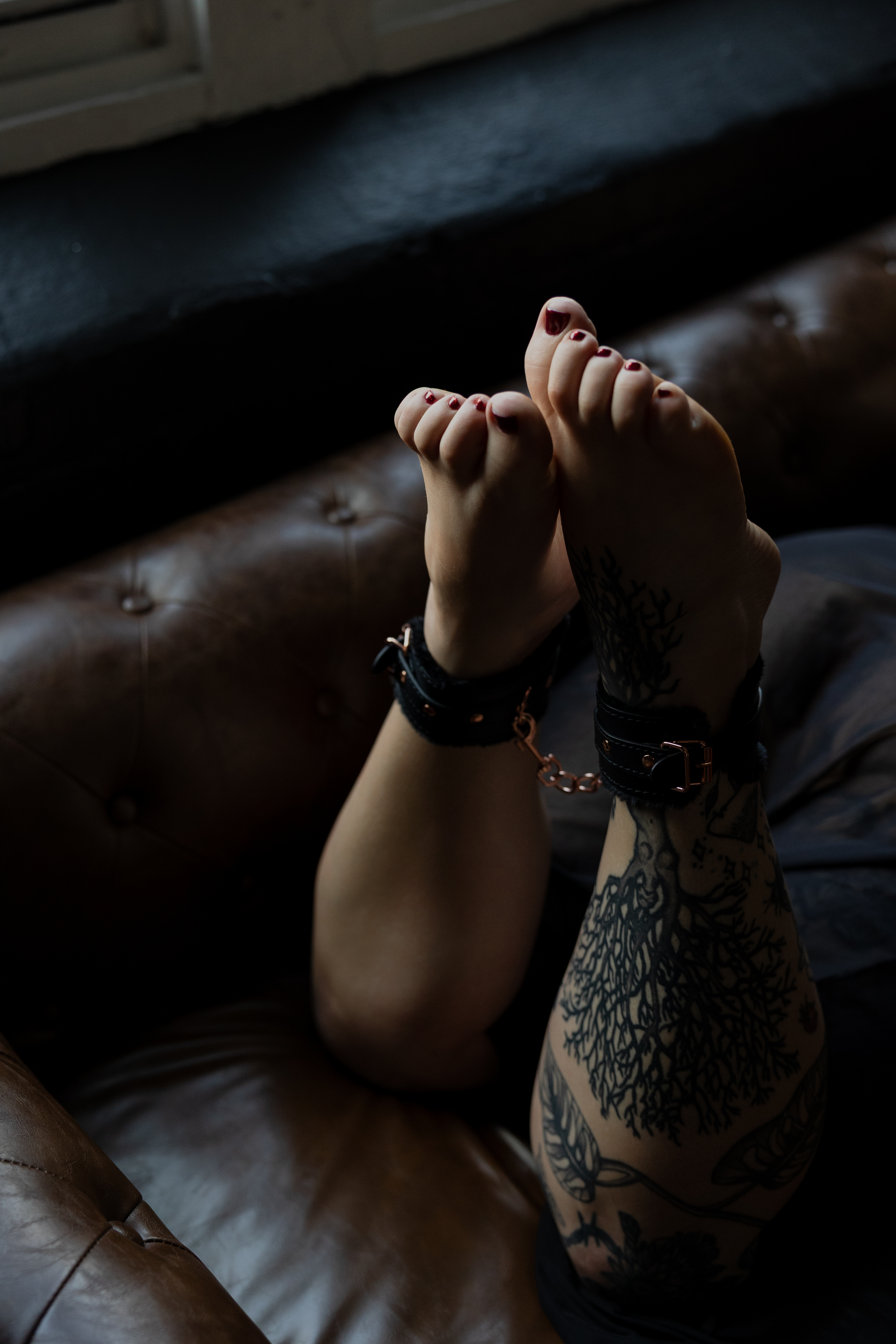 Professional boudoir photography of tattooed legs posed on a leather couch with soft, cinematic window lighting.