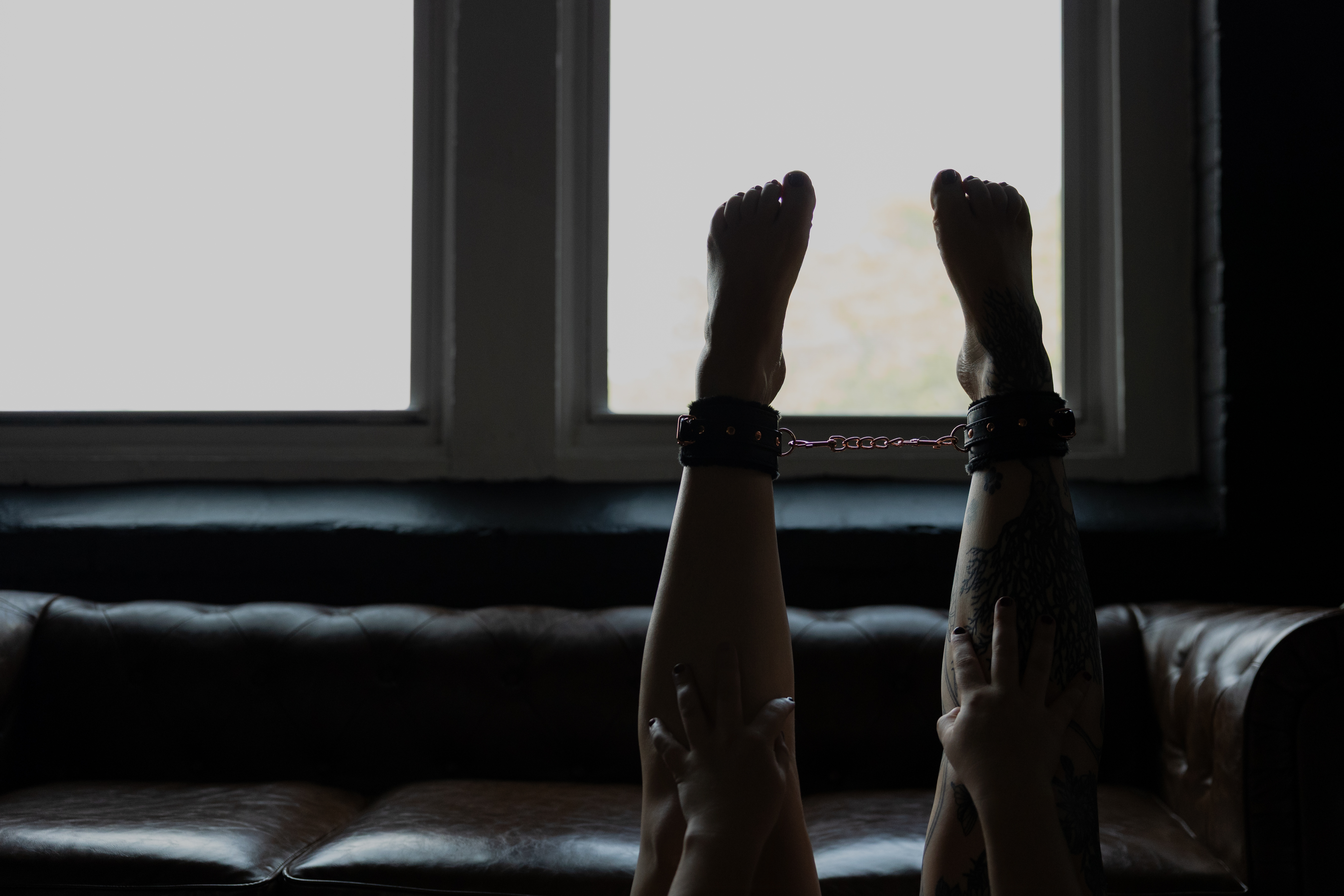 oudoir photograph of tattooed legs resting on a brown leather sofa beneath a window.