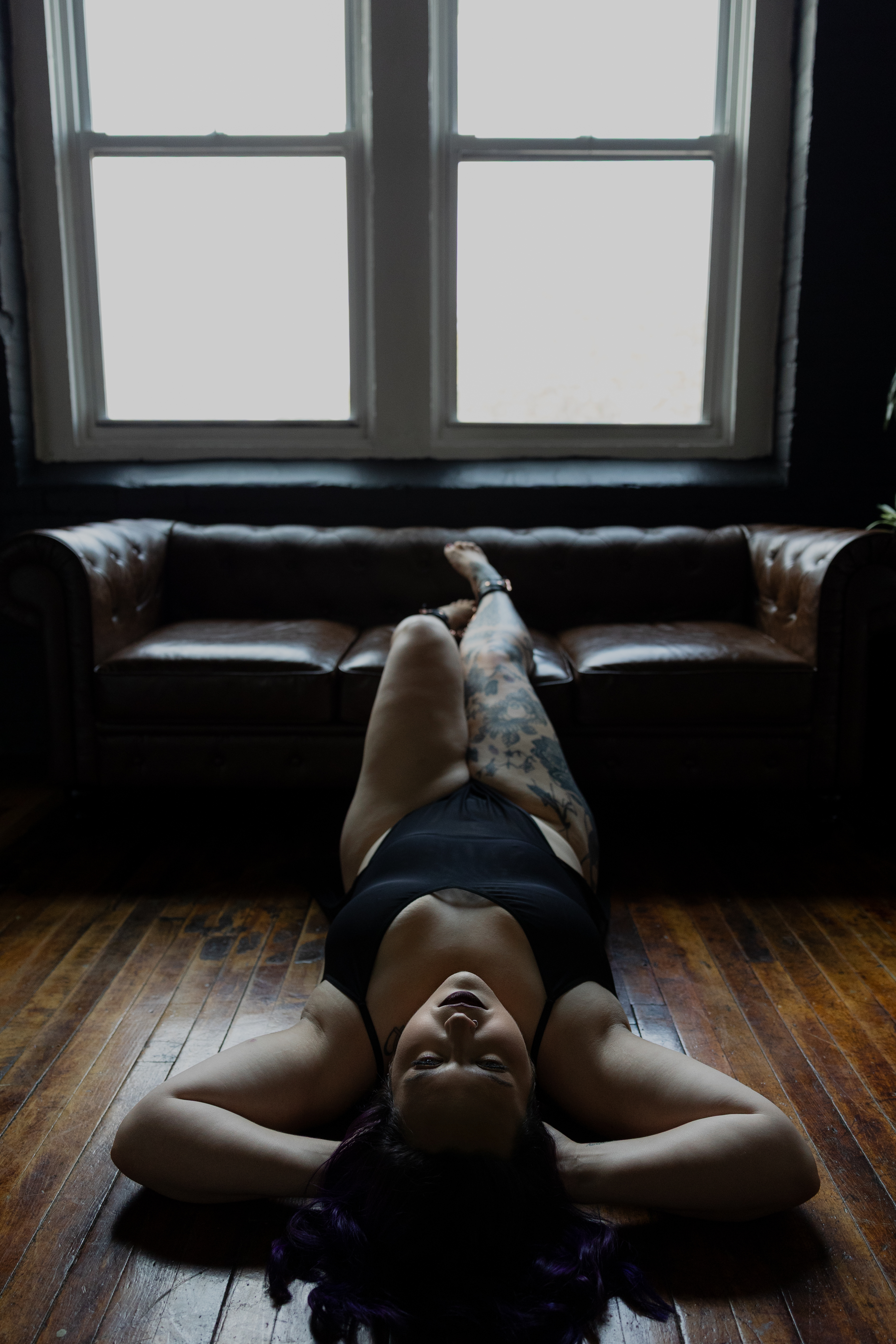 Boudoir photography of a woman in black lingerie lying on a wooden floor beneath large windows, featuring floral leg tattoos and soft natural light.