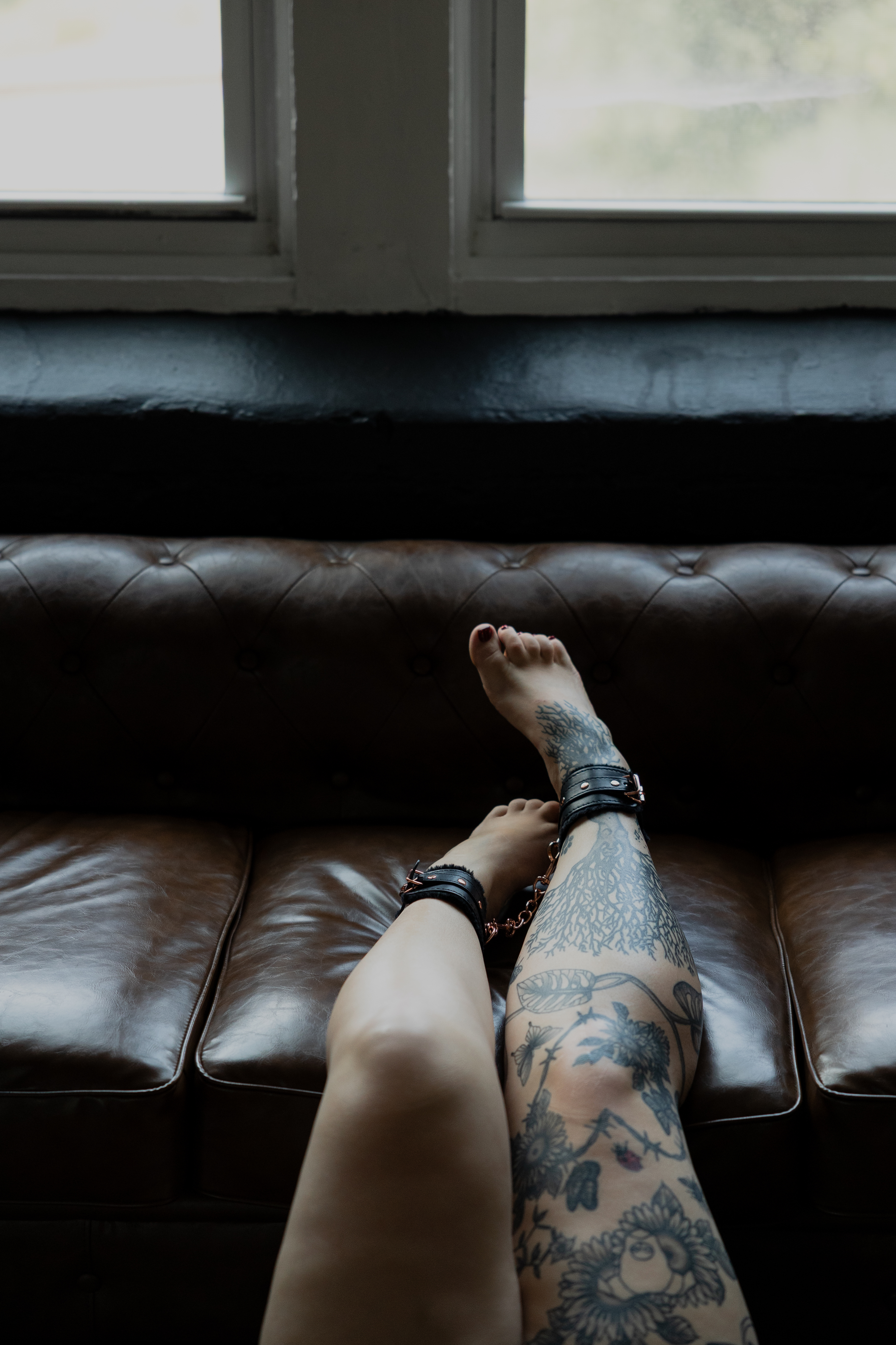 oudoir photograph of tattooed legs resting on a brown leather sofa beneath a window.