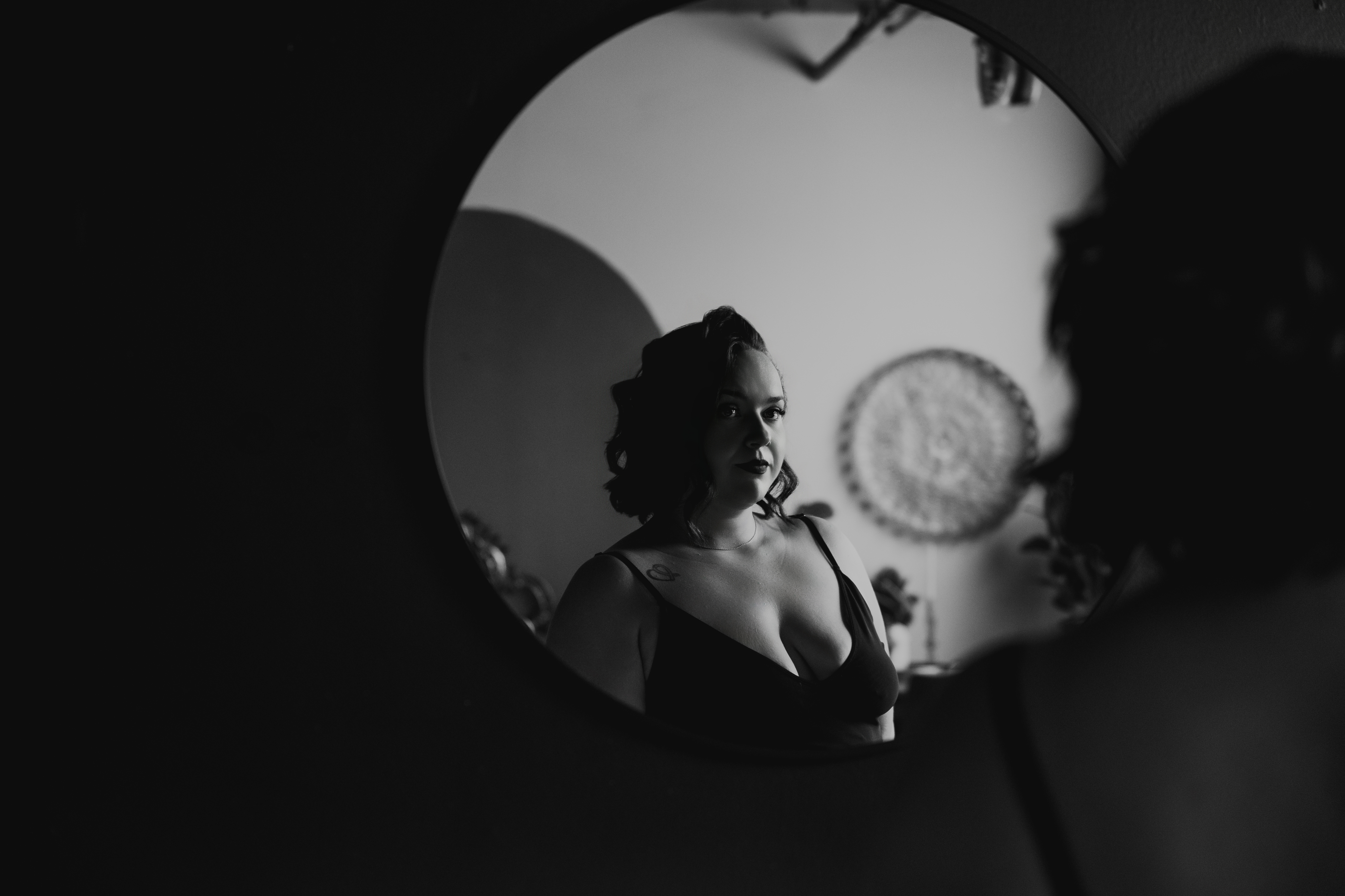 Professional black and white boudoir photography of a woman in lingerie reflected in a round mirror with dramatic lighting.