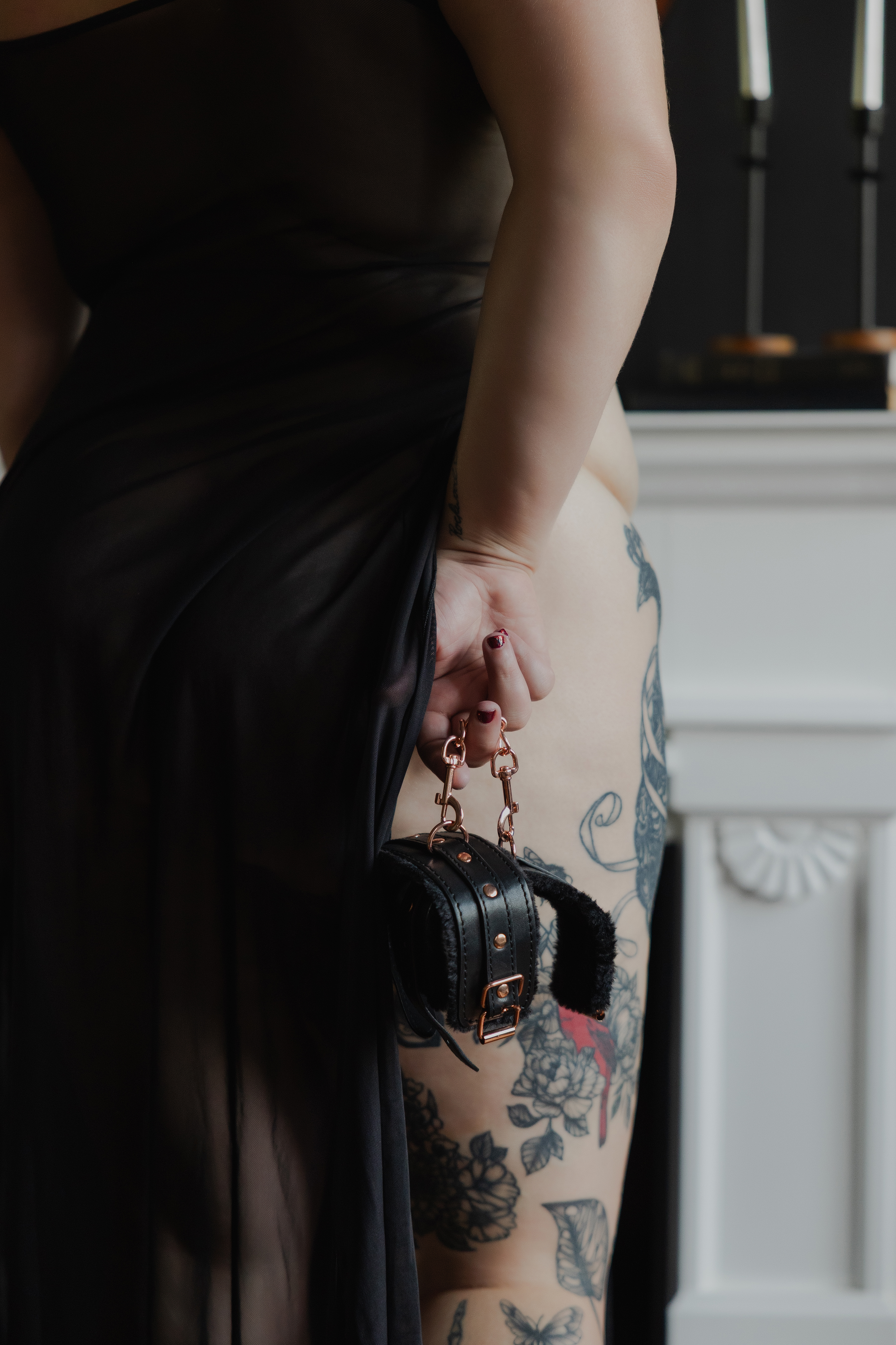Professional boudoir photography close-up capturing styled accessories, black fabric draping, and detailed leg tattoos in natural window light.