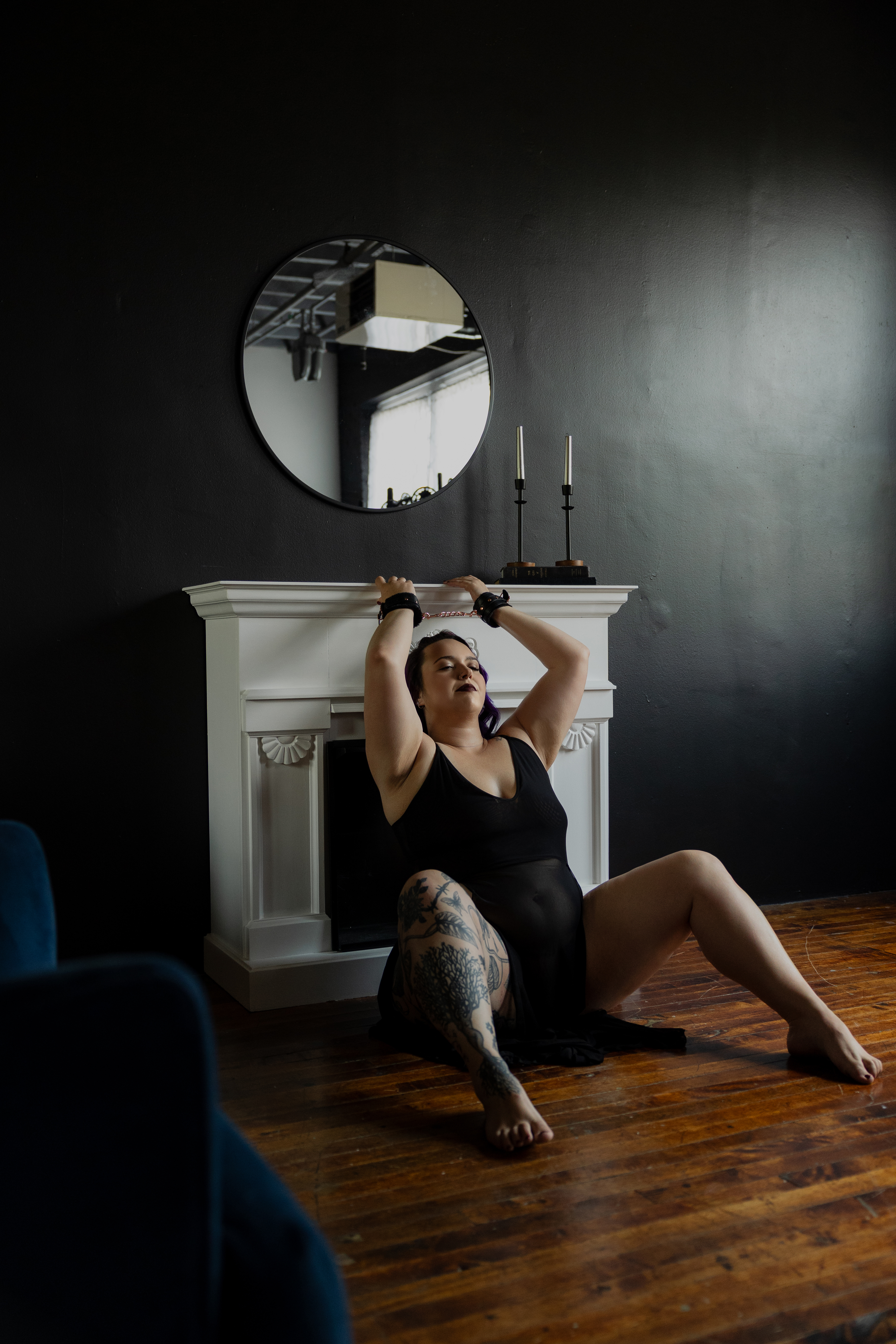 Studio boudoir portrait of a woman in black lingerie sitting in front of a white mantel and round mirror.