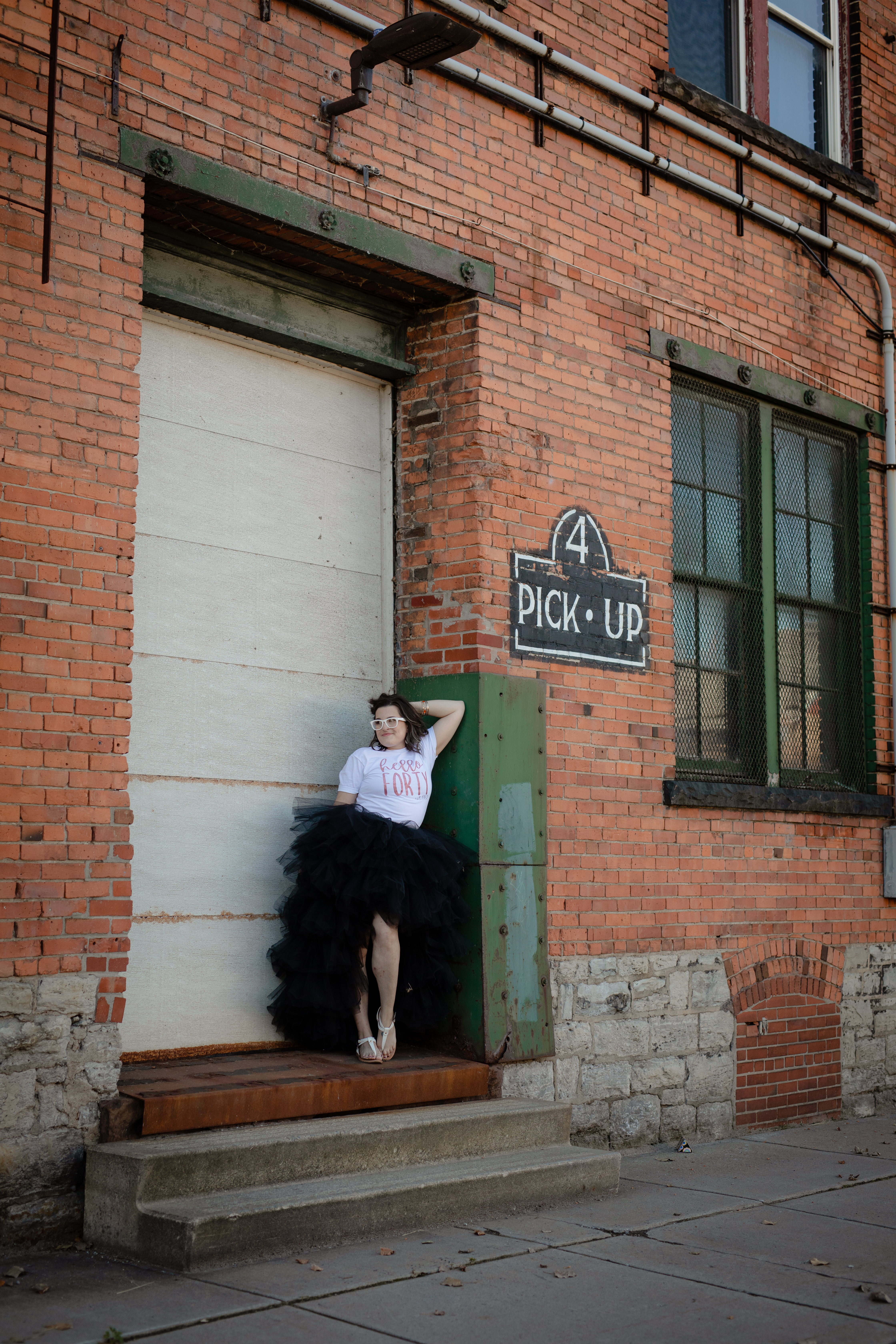 Outdoor urban birthday portrait of a woman in a “Hello Forty” shirt posing near a brick building and garage door. Outdoor urban birthday portrait of a woman in a “Hello Forty” shirt posing near a brick building and garage door.