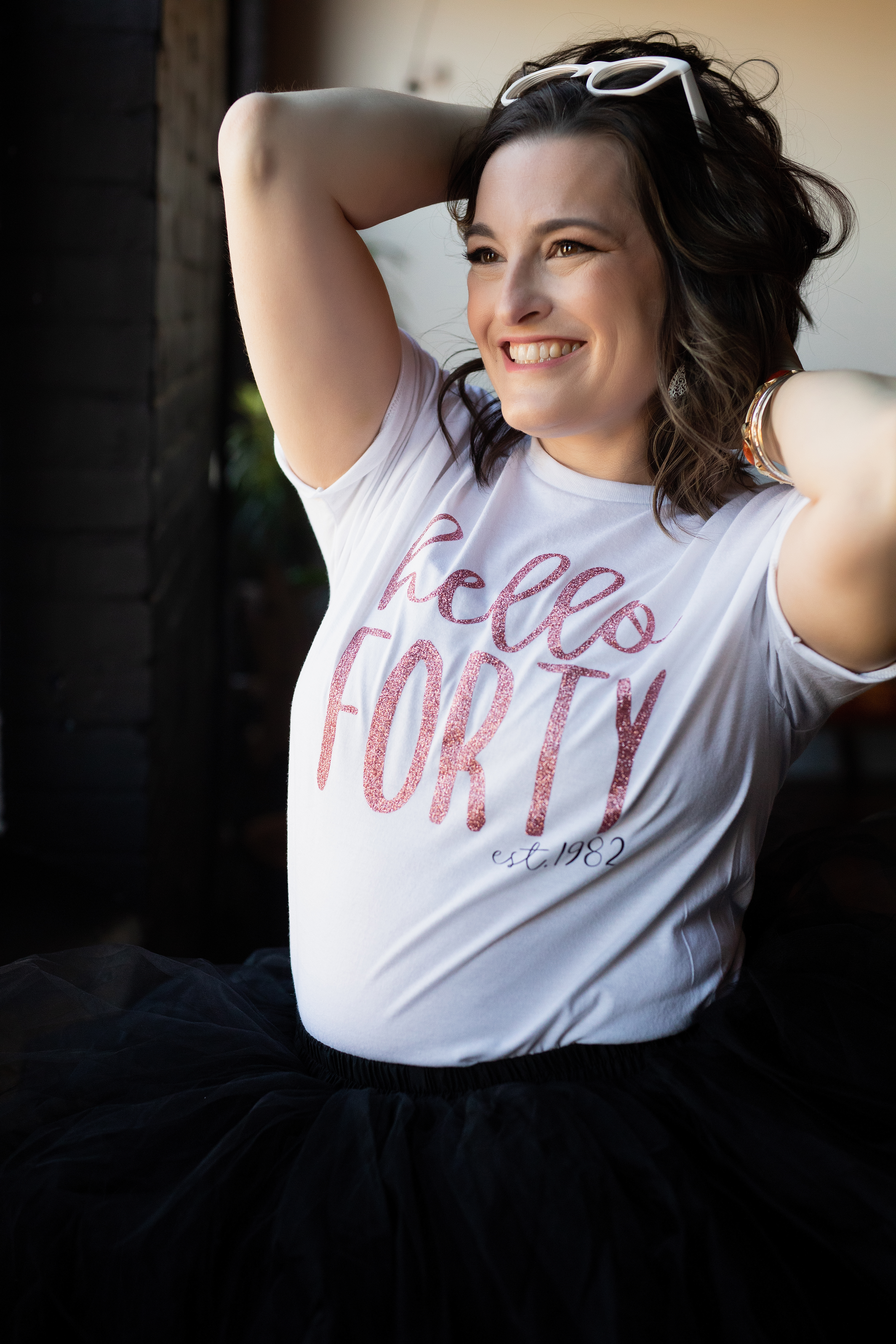 Lifestyle portrait of a woman wearing a “Hello Forty” birthday shirt, smiling with arms raised in natural light. Lifestyle portrait of a woman wearing a “Hello Forty” birthday shirt, smiling with arms raised in natural light.