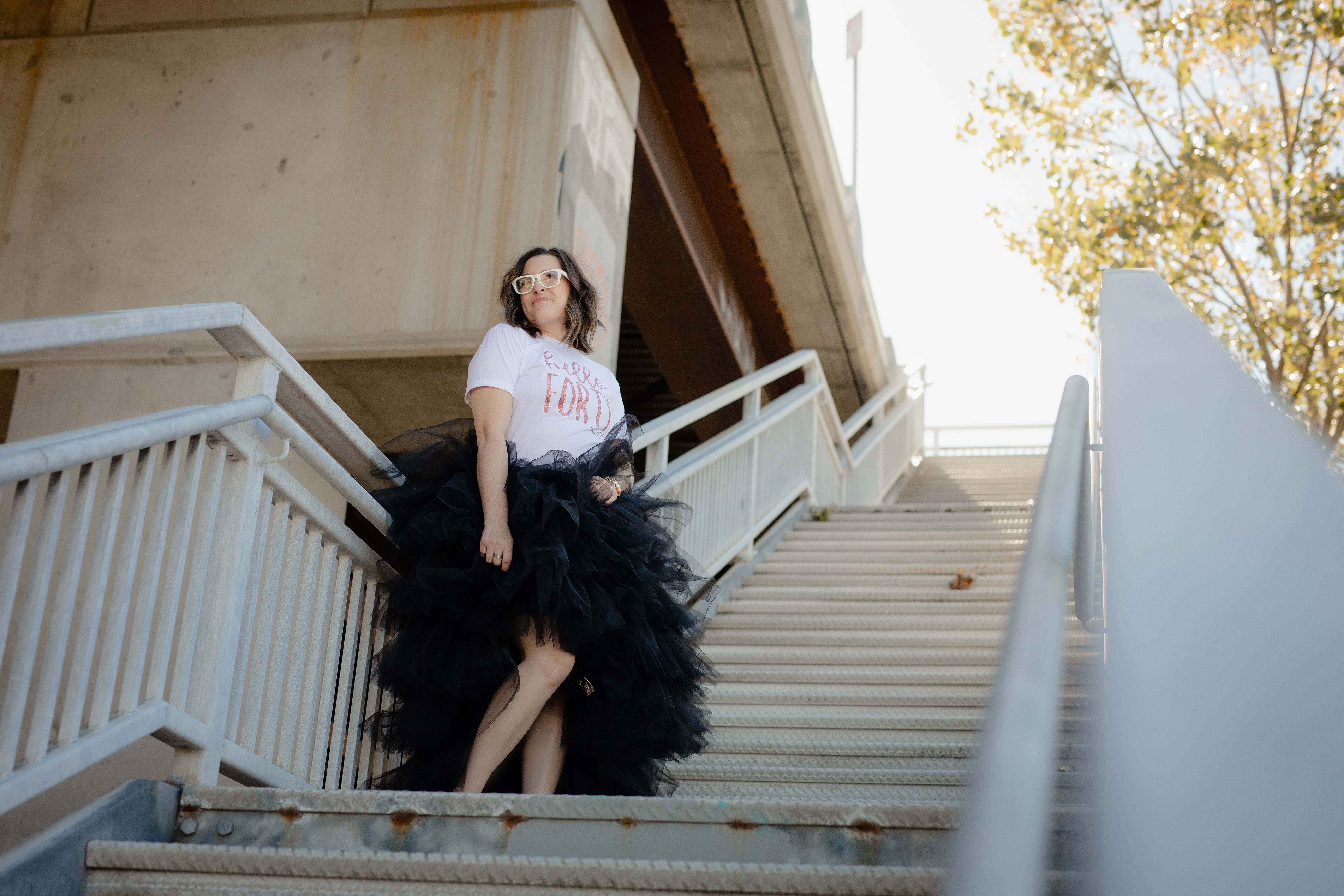 Woman celebrating her 40th birthday posing on a staircase in a white “Hello Forty” shirt and black tulle skirt. Woman celebrating her 40th birthday posing on a staircase in a white “Hello Forty” shirt and black tulle skirt.
