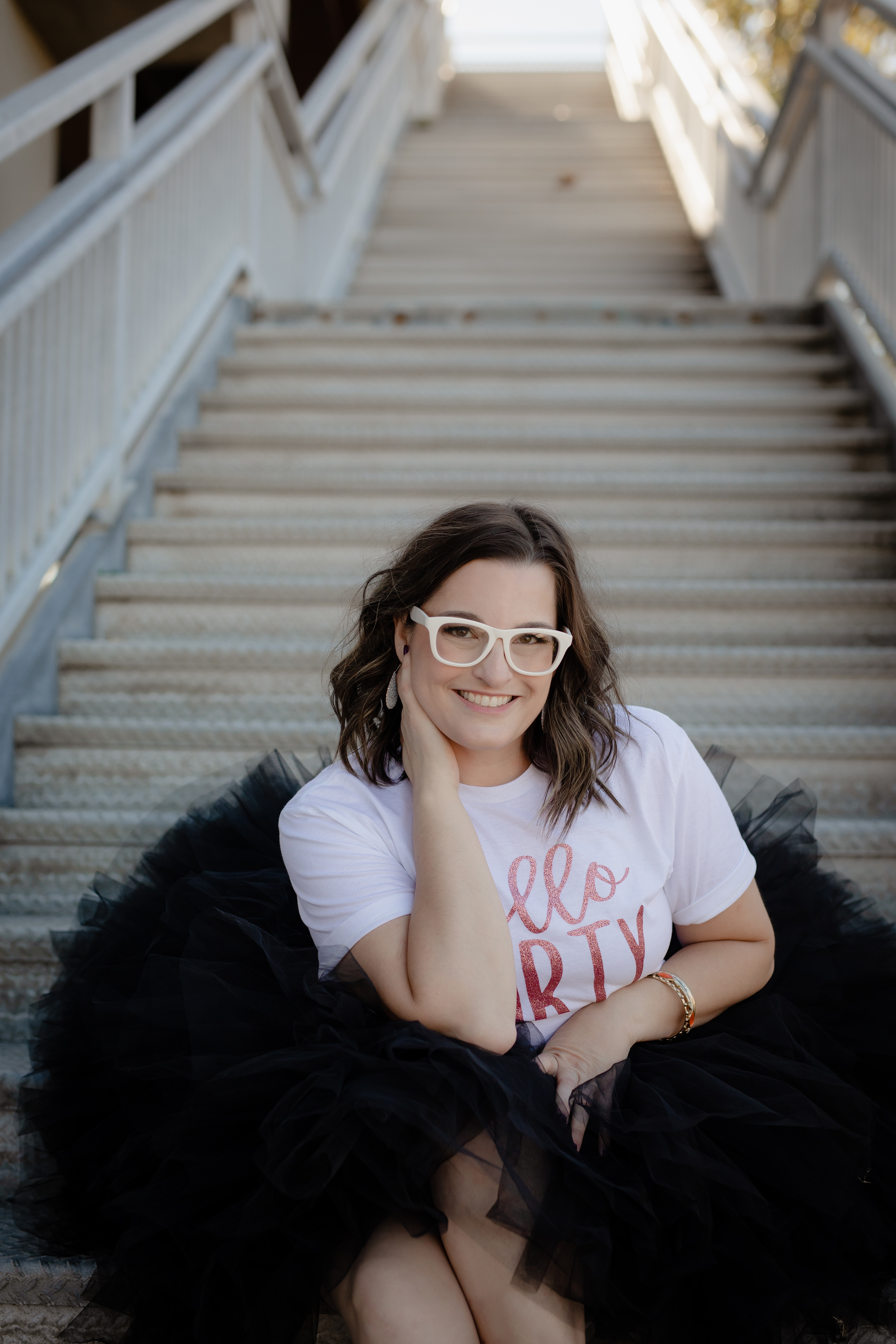 Smiling woman in a “Hello Forty” t-shirt sitting on outdoor stairs during a 40th birthday portrait session. Smiling woman in a “Hello Forty” t-shirt sitting on outdoor stairs during a 40th birthday portrait session.