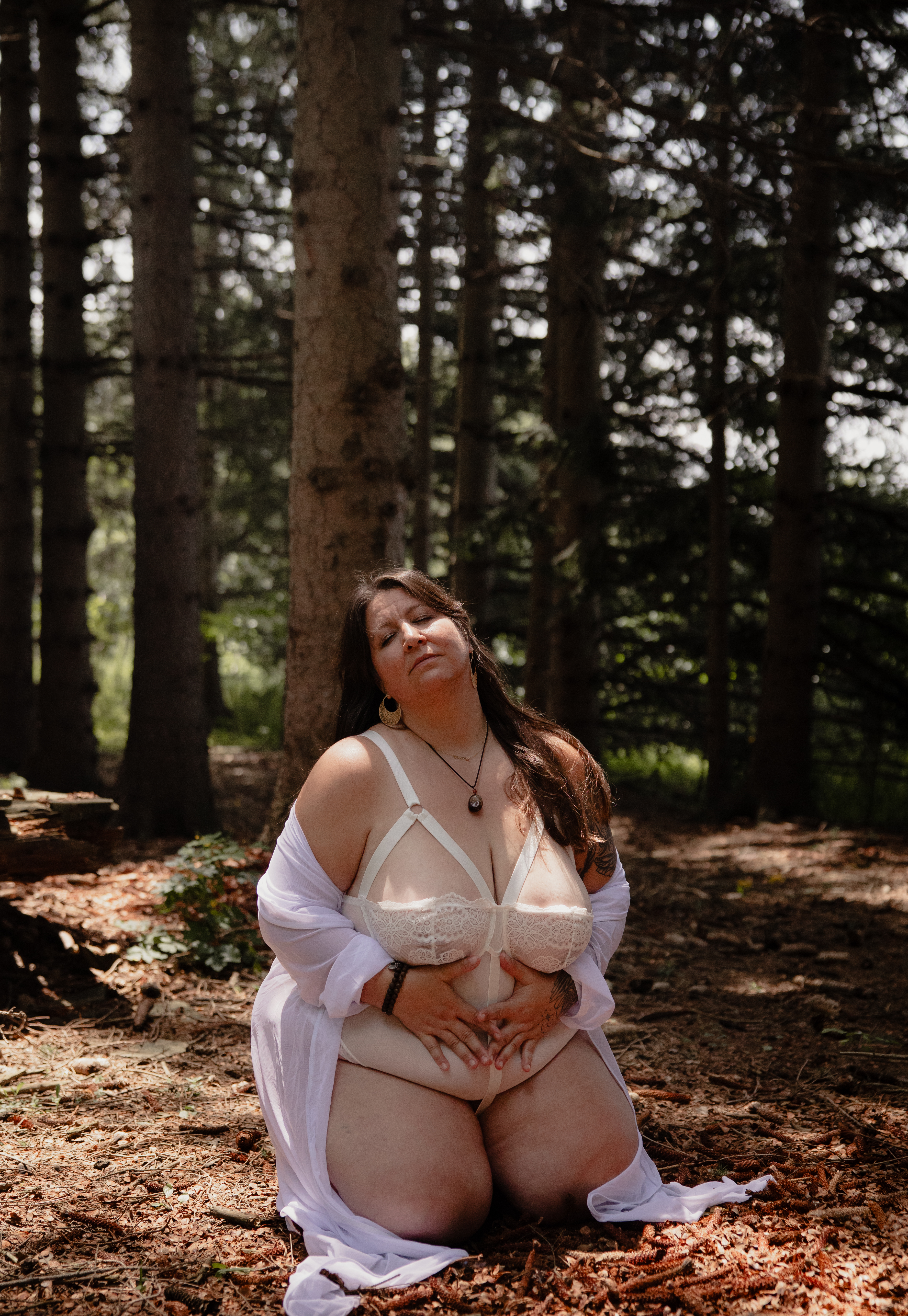 Woman twirling in a white dress in a pine forest during a boudoir photography session.