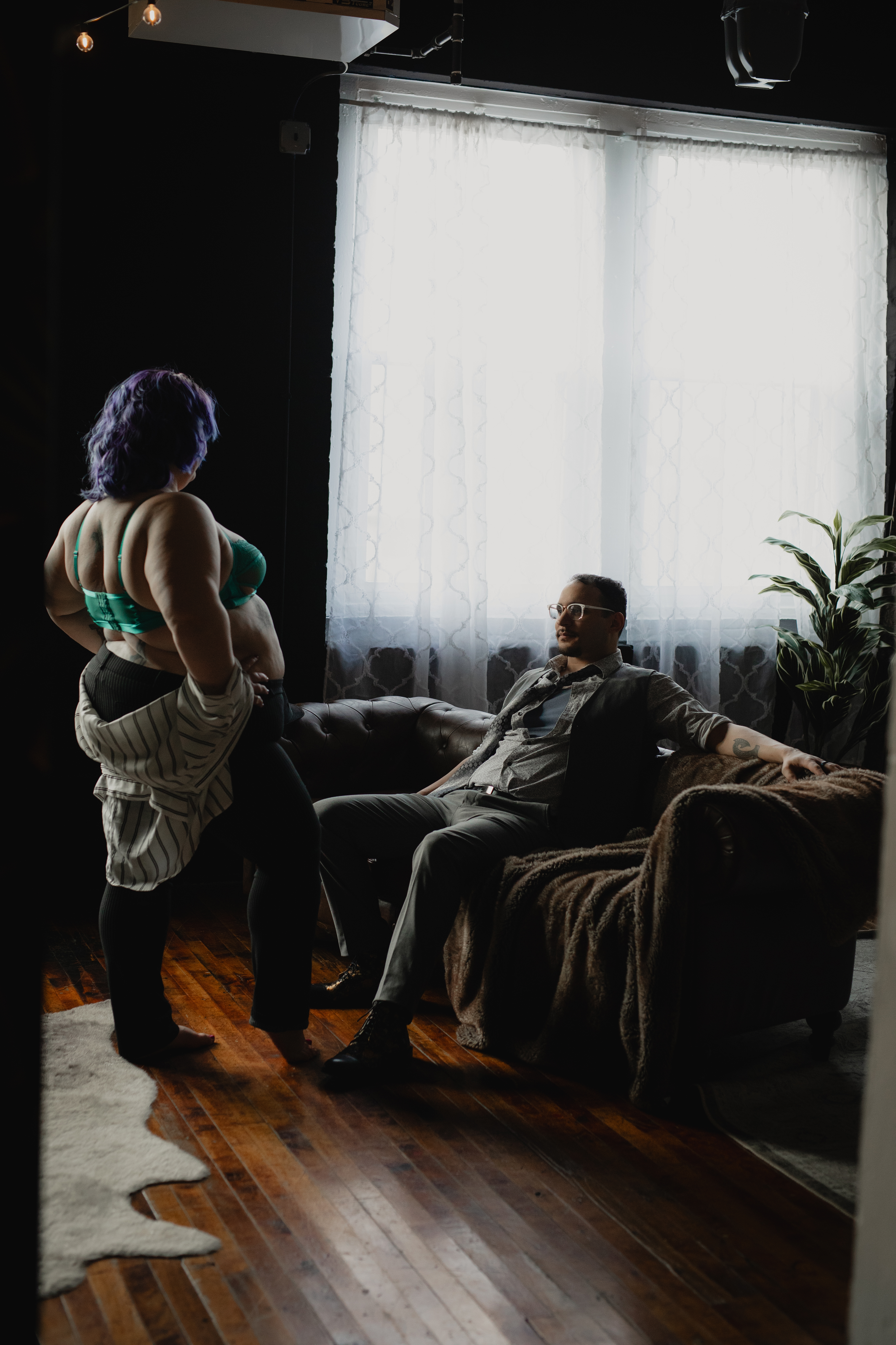 Woman standing in lingerie facing partner seated on a couch in a boudoir session.