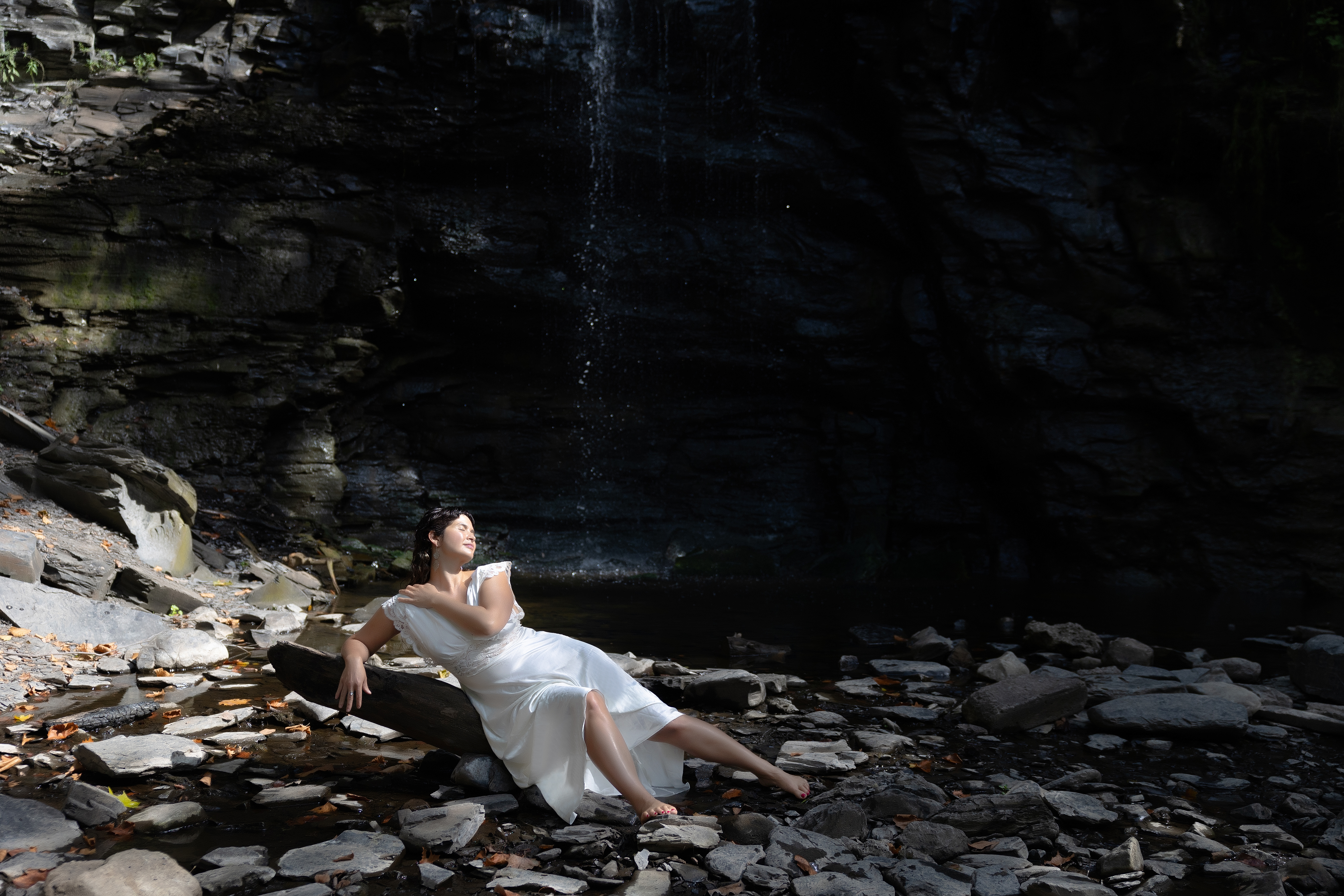 Outdoor boudoir portrait of a woman sitting by a waterfall in a natural setting.