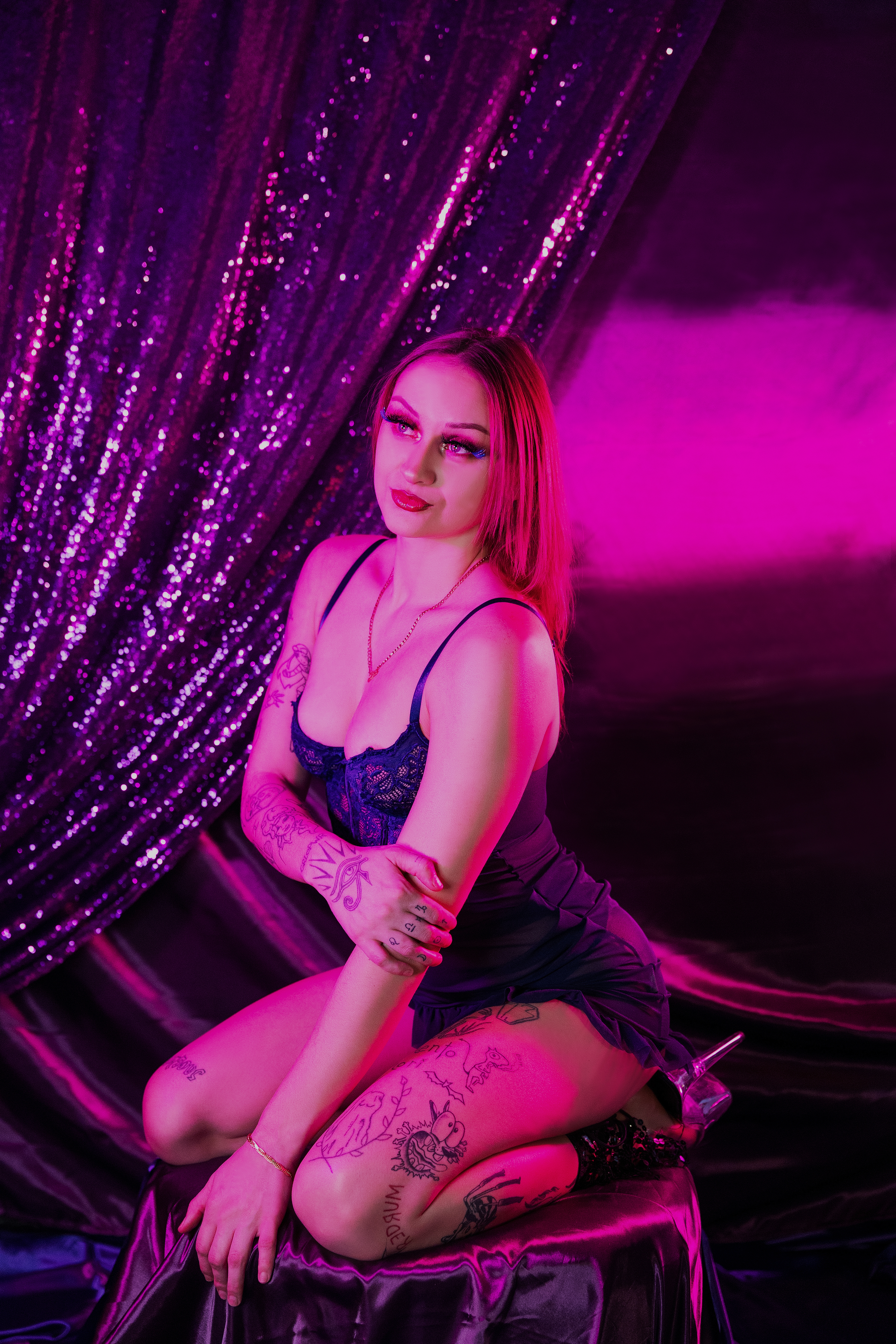 Woman in lingerie standing in front of shimmering curtains in a neon boudoir photoshoot.