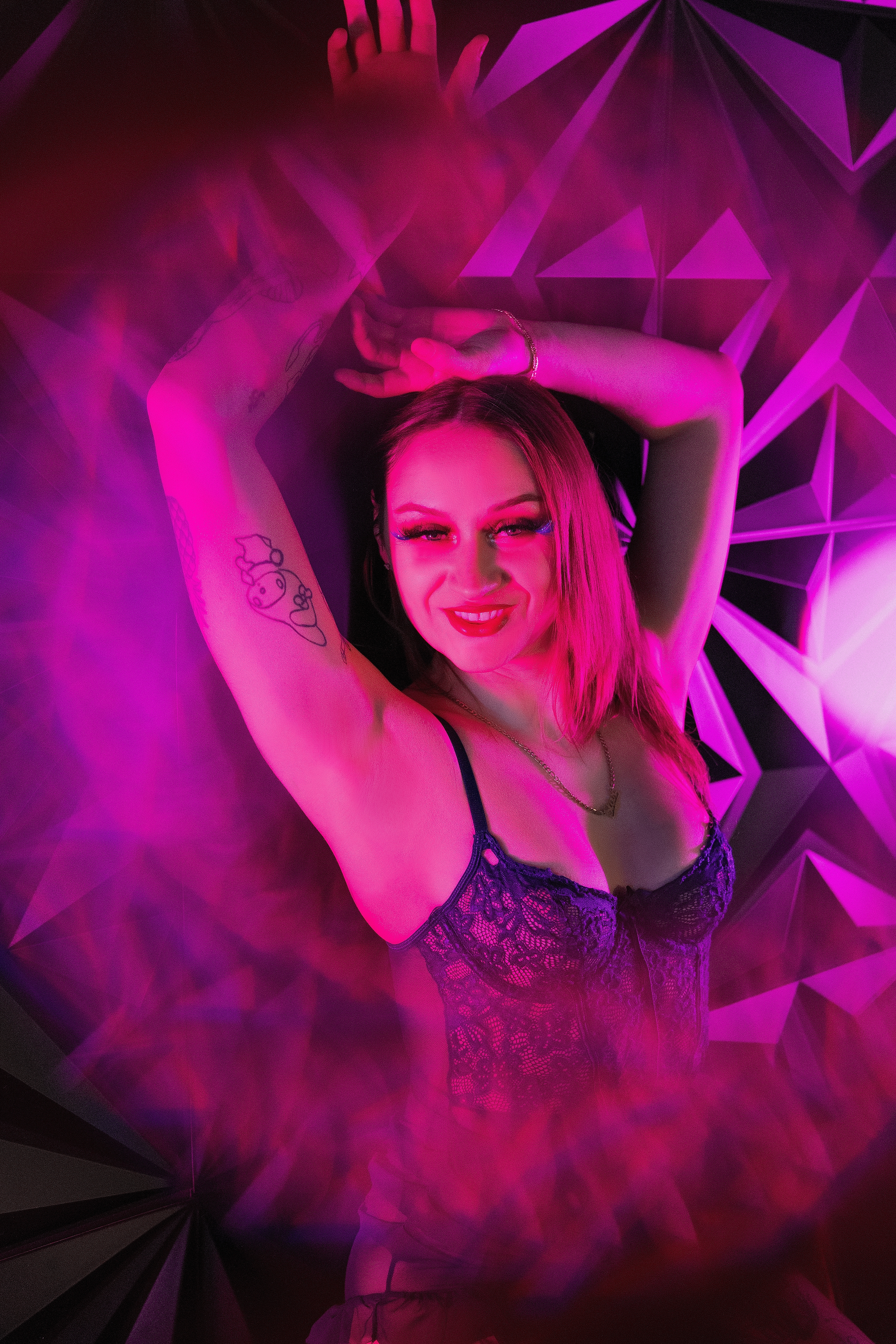 Woman posing with raised arms in a dramatic neon boudoir photography setup.