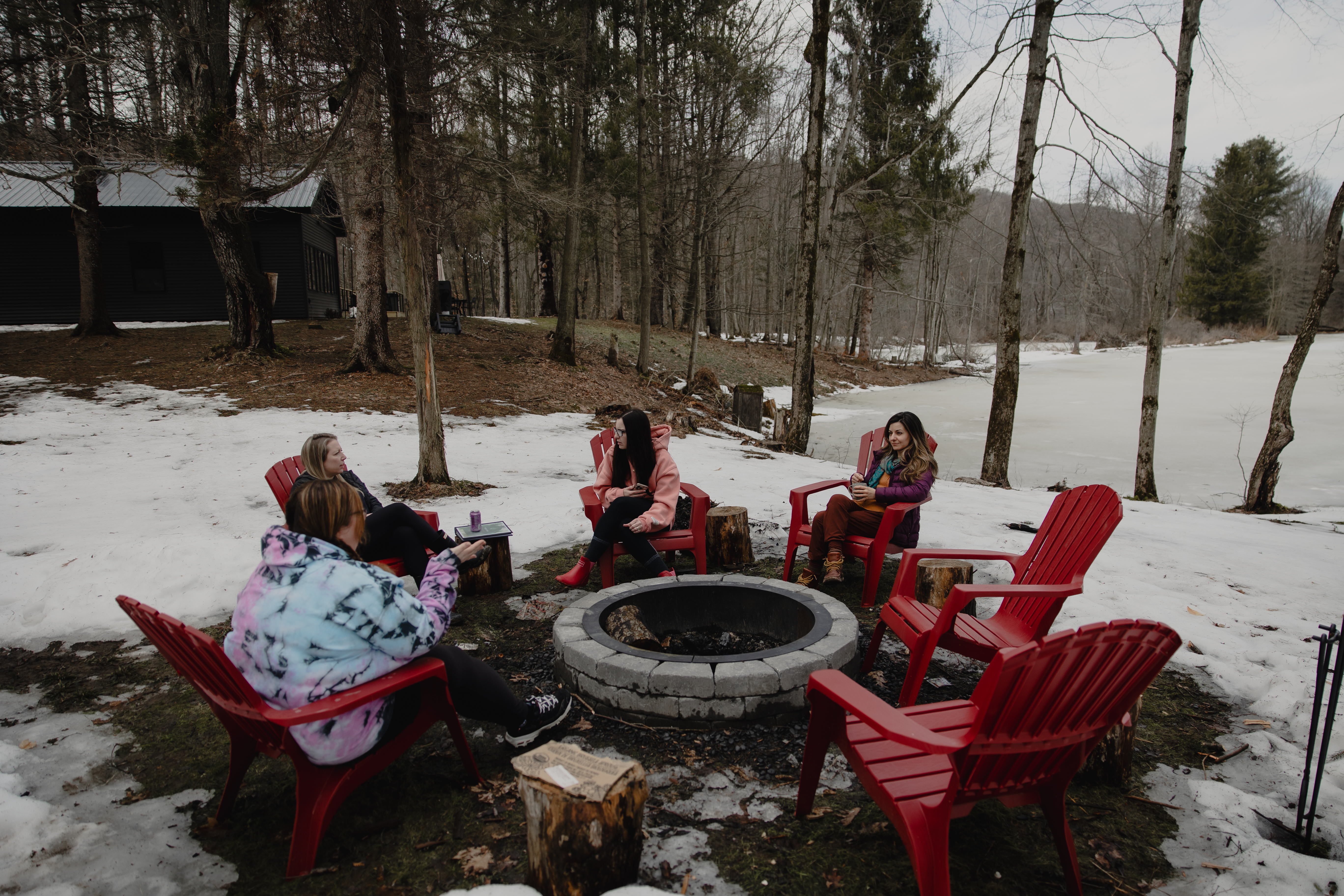 Group of women gathered outdoors around a fire pit in a wooded retreat setting.