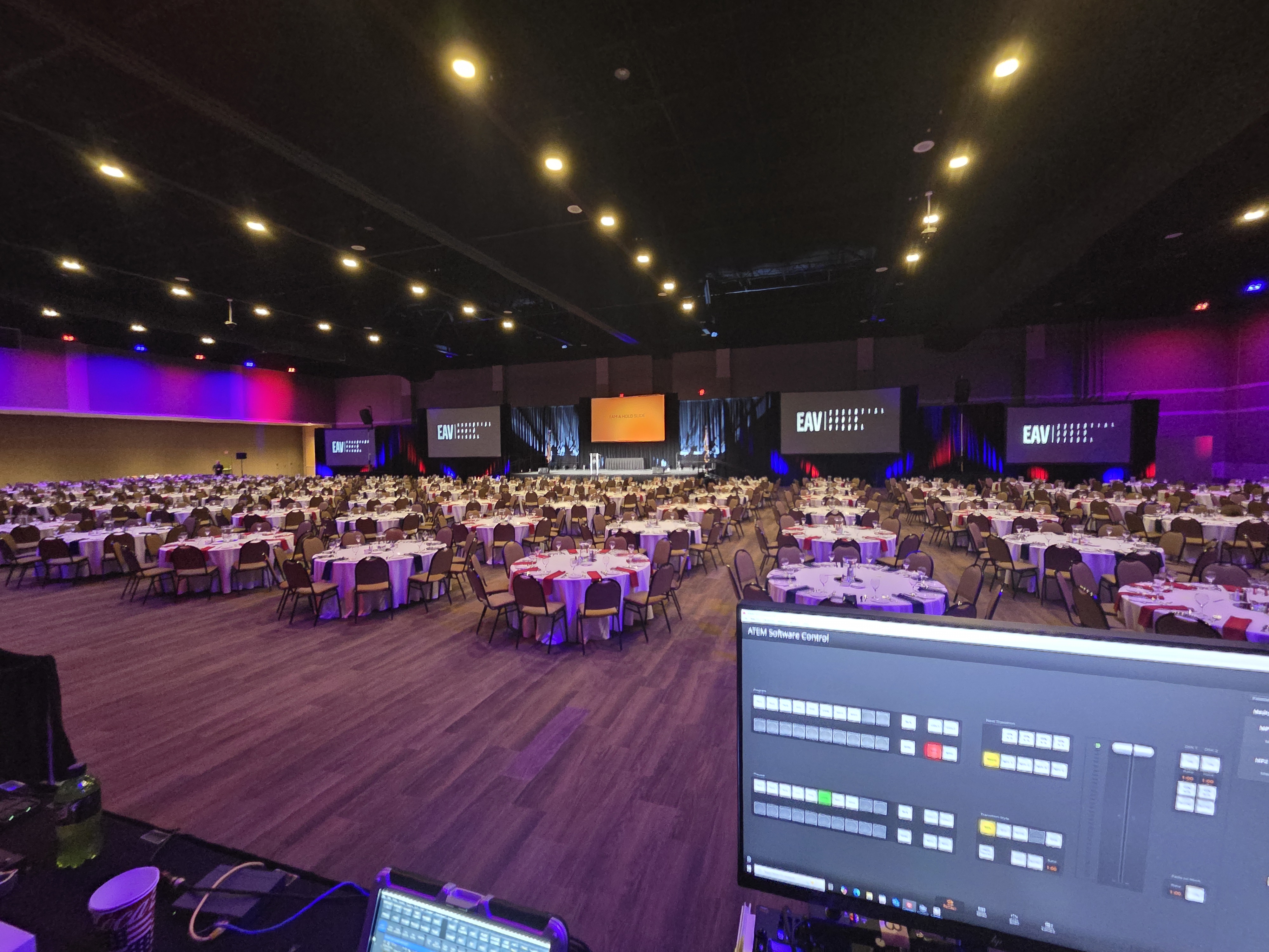 An Essential AV gala general session — a full ballroom of round tables and multiple screens under warm and purple lighting, with the front-of-house console in the foreground.