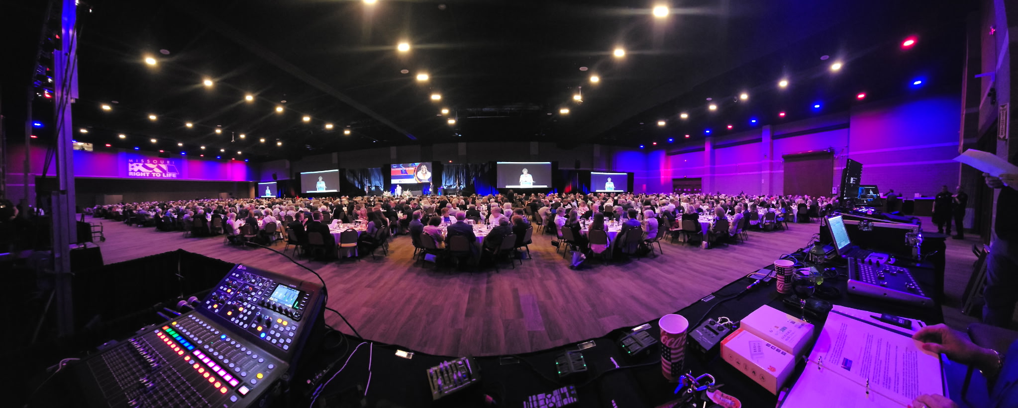 An Essential AV convention general session seen from the front-of-house console — a full hall of round tables facing a wide stage and screen.
