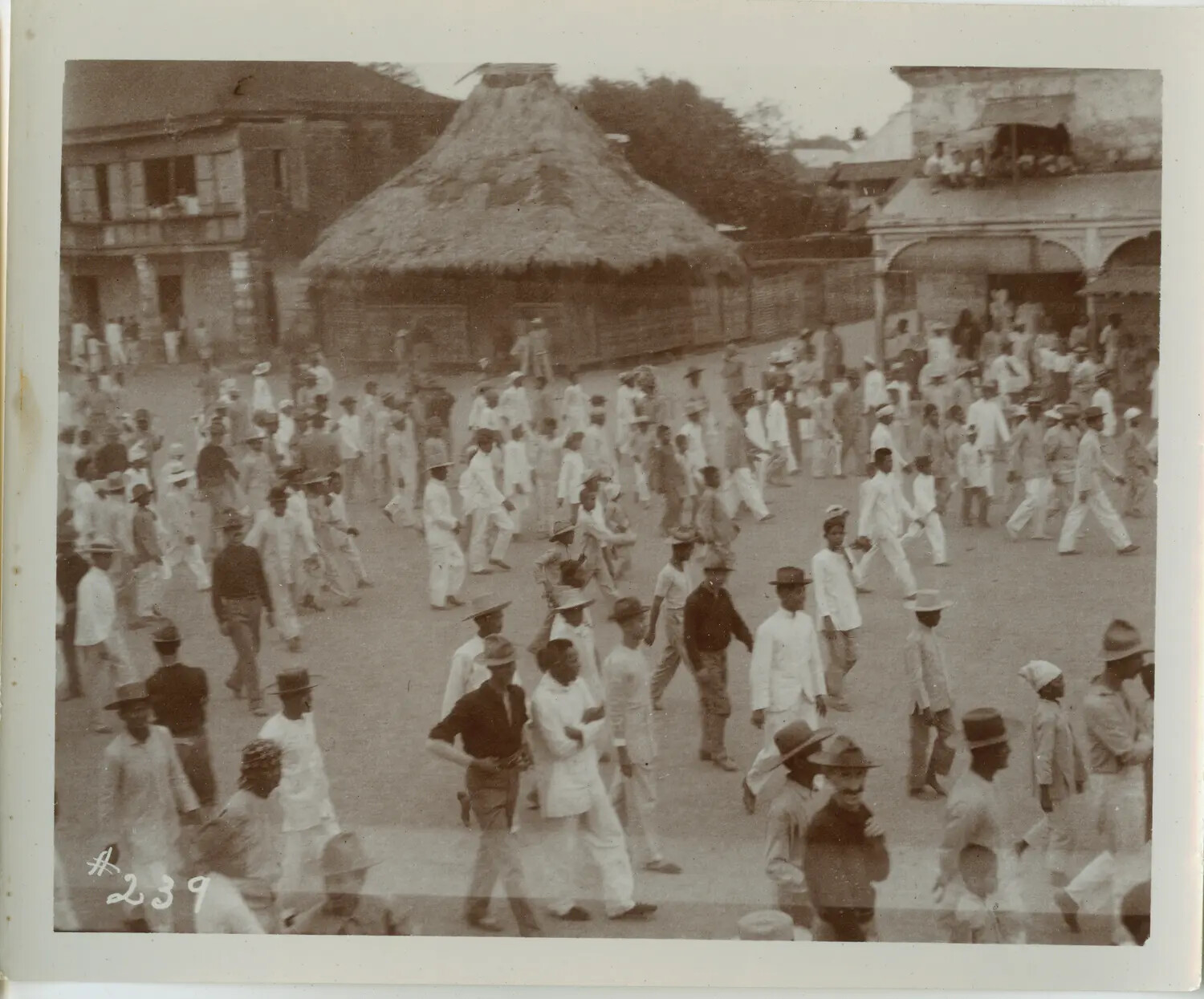 Crowd of Filipino Men in Town Plaza Beside Conical Thatched Structure, Philippines, Early 1900s