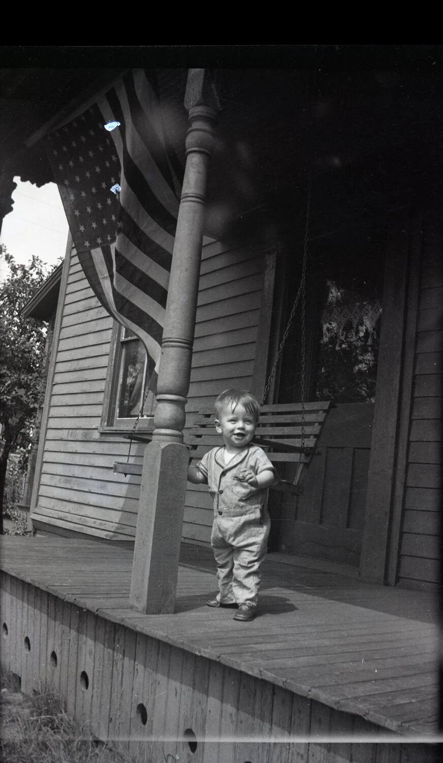 Toddler Standing on a Porch Beneath a Hanging American Flag