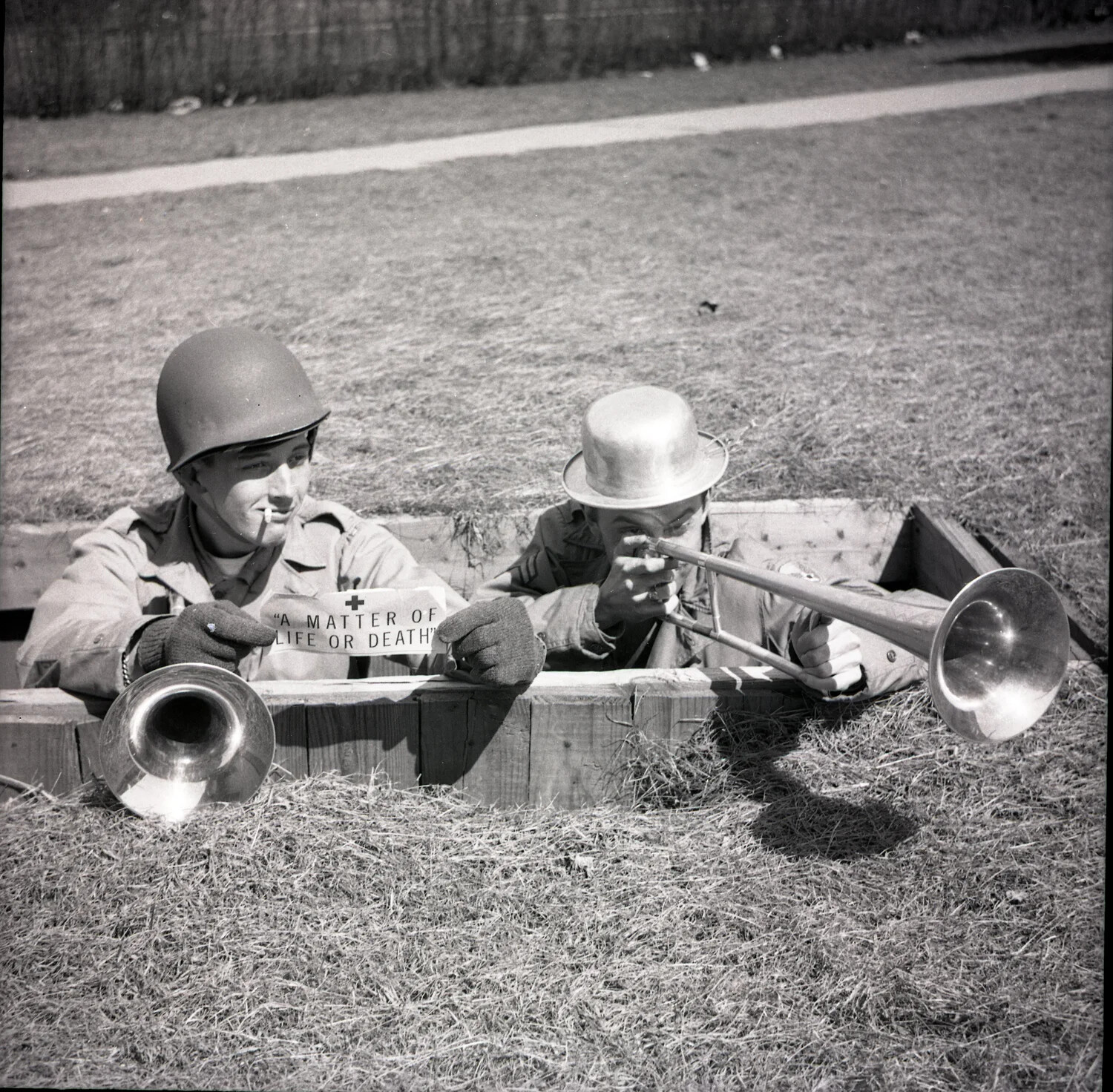 Soldiers in Mock Foxhole with Trombone and Red Cross Placard, "A Matter of Life or Death"