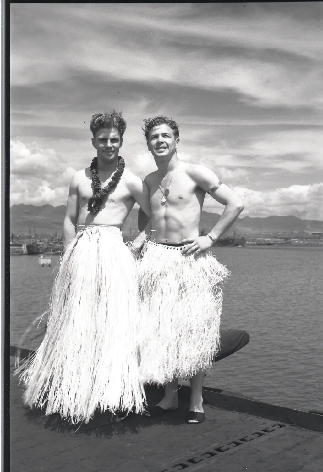 Two U.S. Navy Sailors in Grass Skirts on Ship's Deck, Hawaiian Harbor, WWII
