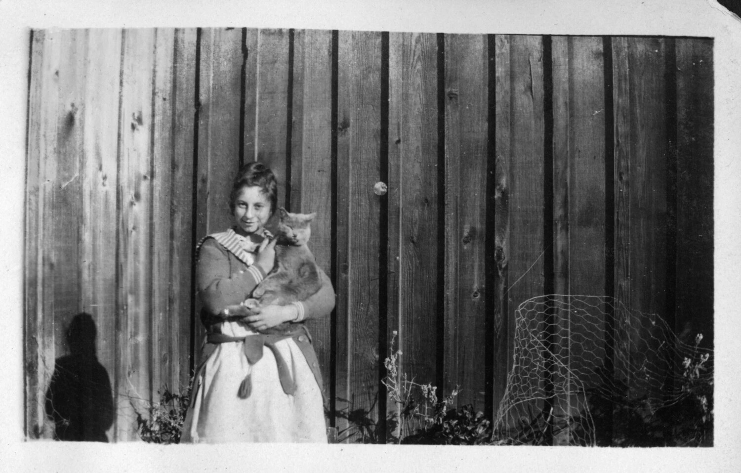 Girl in Sailor Dress Holding Cat Against Wood Fence, c. 1910s–1920s