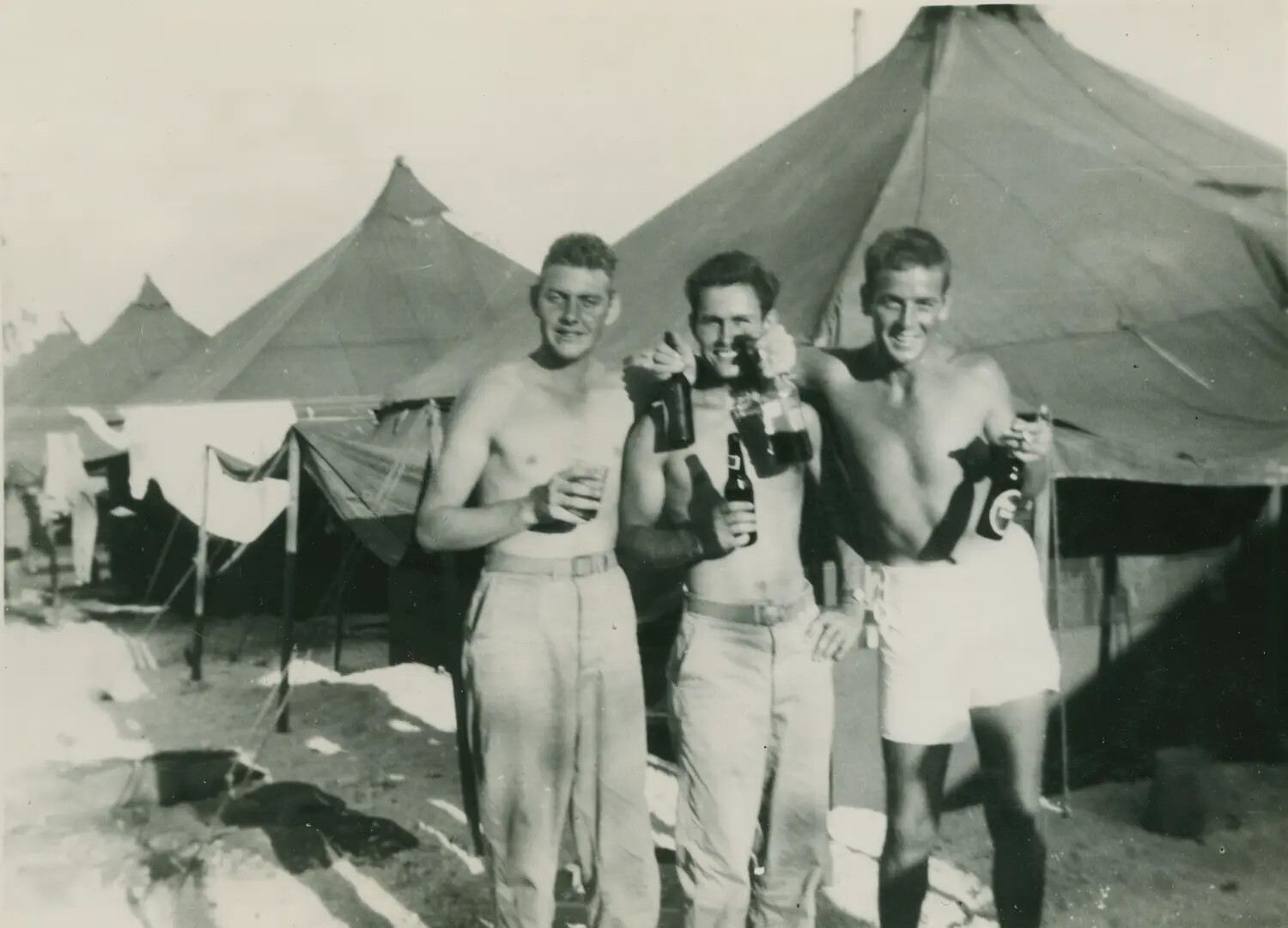 Three Servicemen Toasting with Beer Bottles at a Tent Camp, WWII Era
