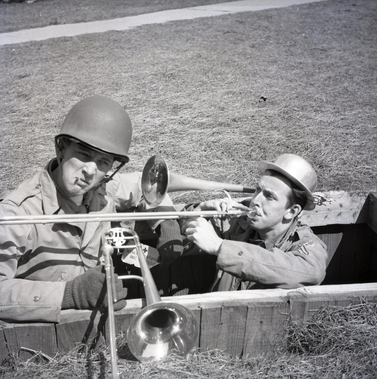 Military Bandsmen Staging Comic Trombone Duet in Mock Foxhole, with Red Cross Appeal Poster