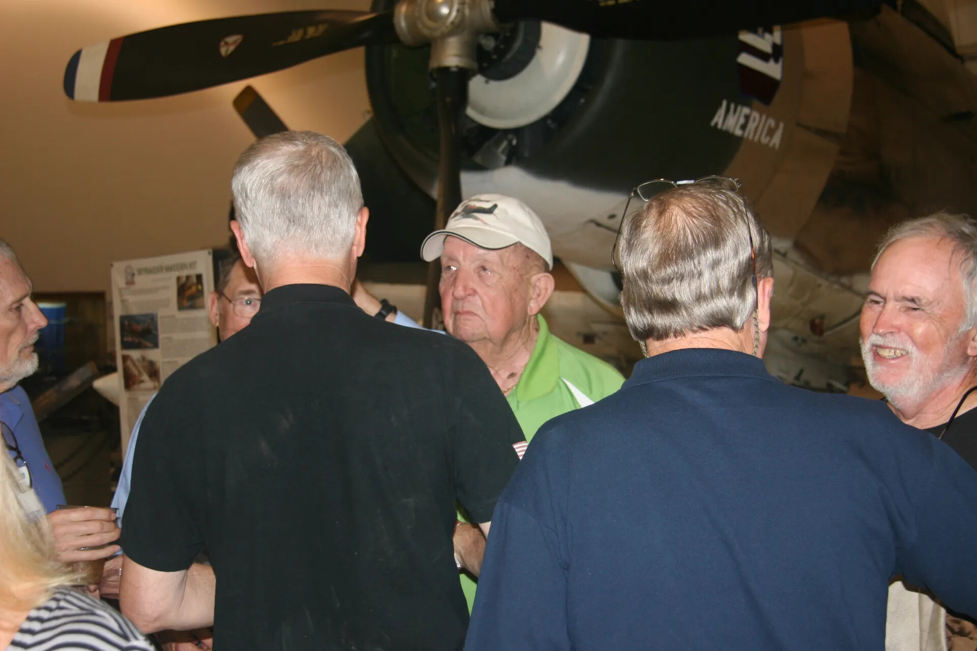 Reunion Attendees Conversing Beneath Aircraft Nose Marked 'AMERICA', Museum Hangar