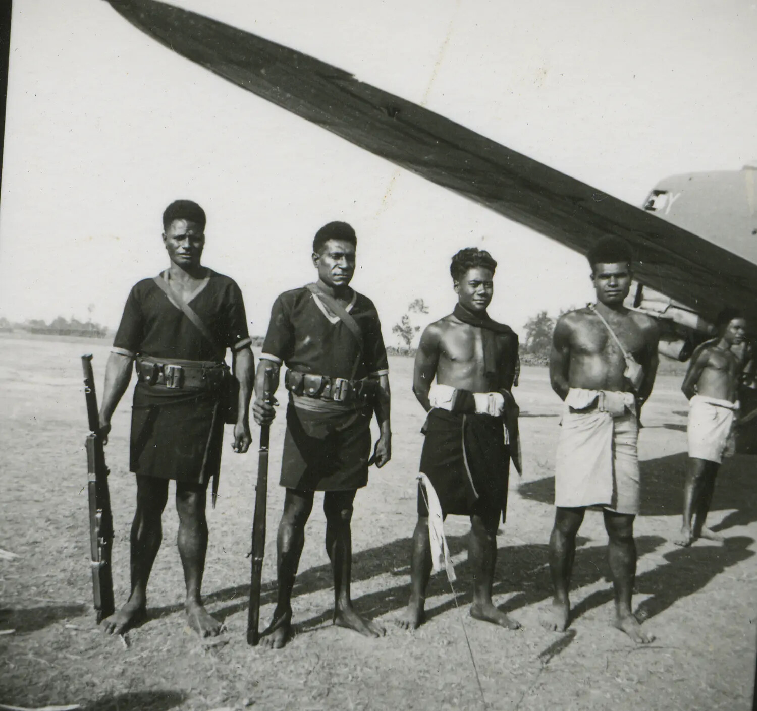 Armed New Guinean Constabulary Members and Local Men Beneath Aircraft Wing, World War II Era