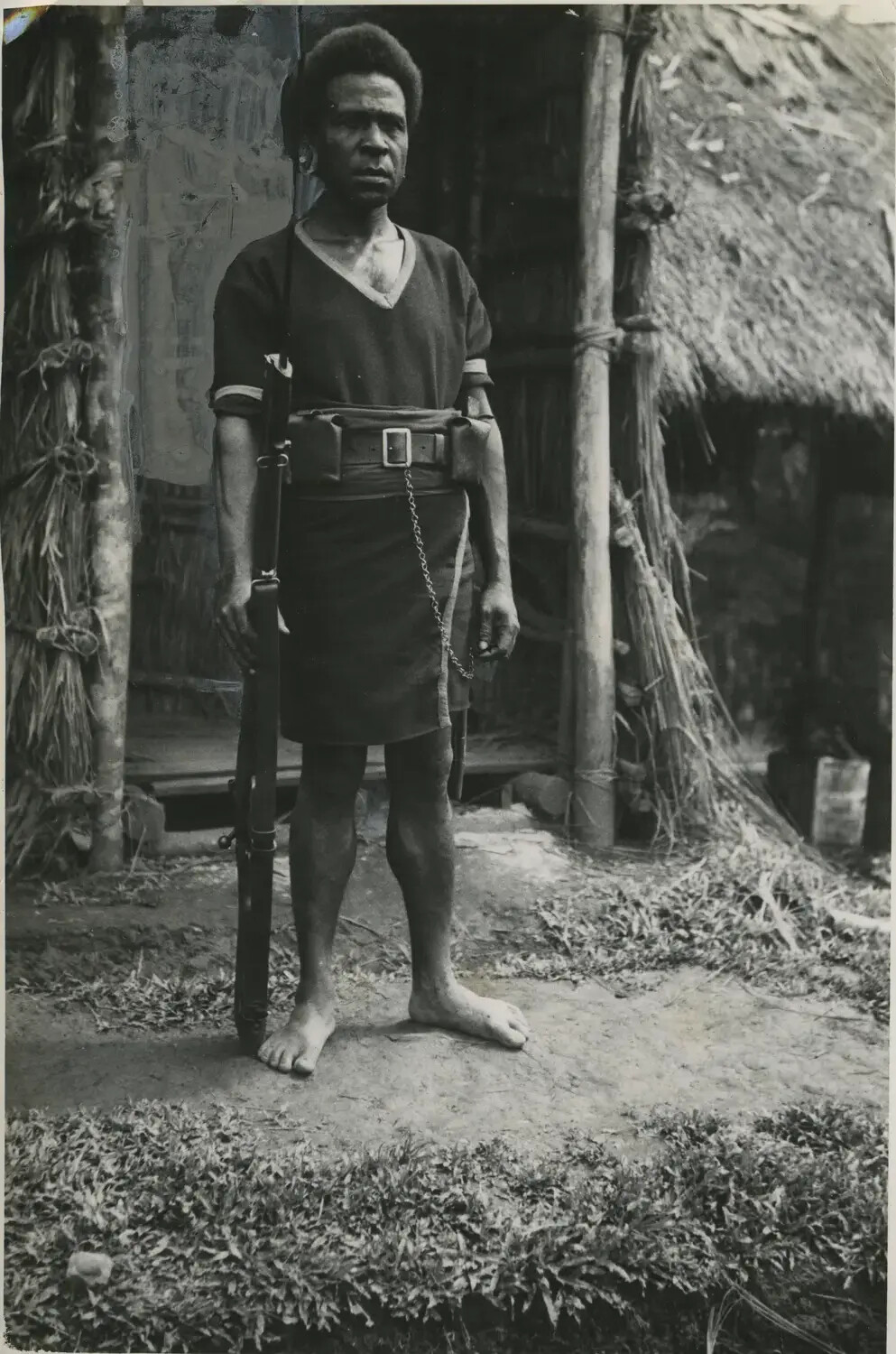 Armed Papua New Guinean Man in Traditional Dress Near Thatched Dwelling, Southwest Pacific, WWII Era