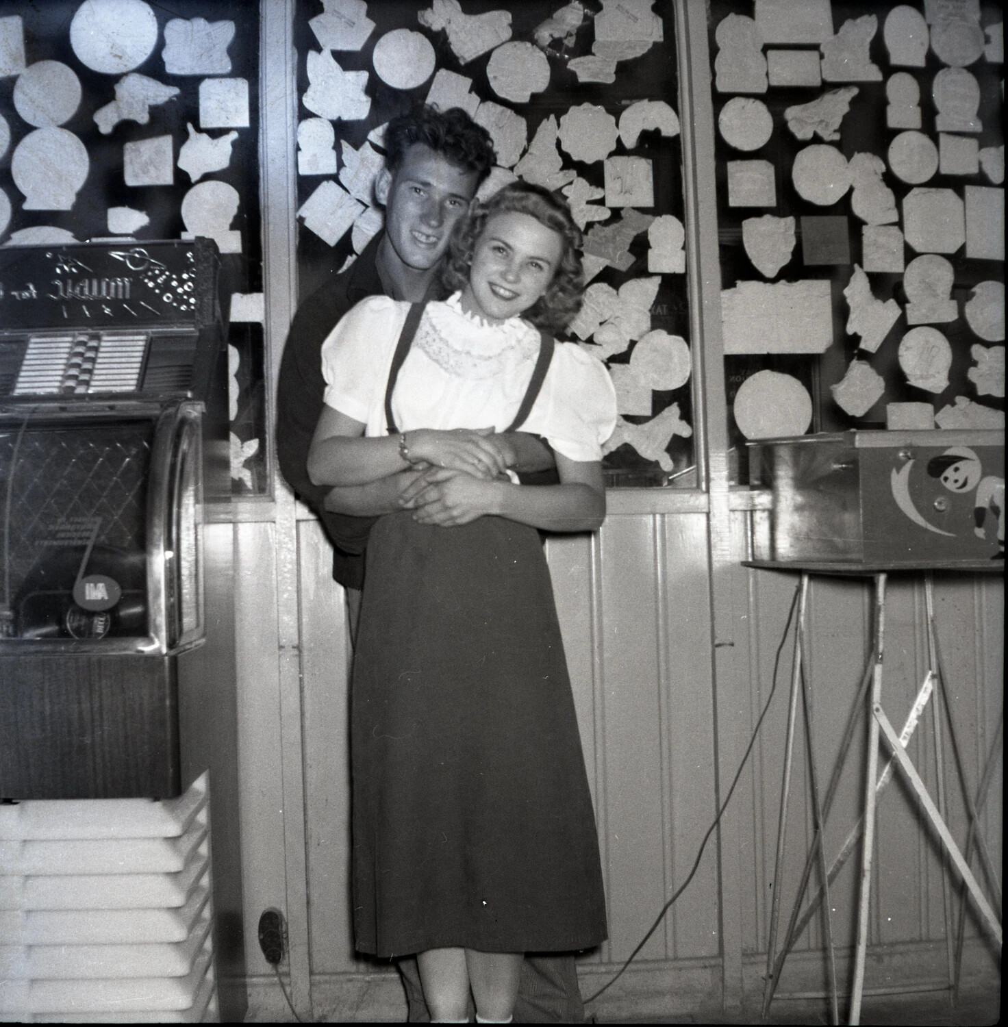 Young Couple Embracing in a Boardwalk Arcade Beside a Jukebox and Plaster Novelty Display