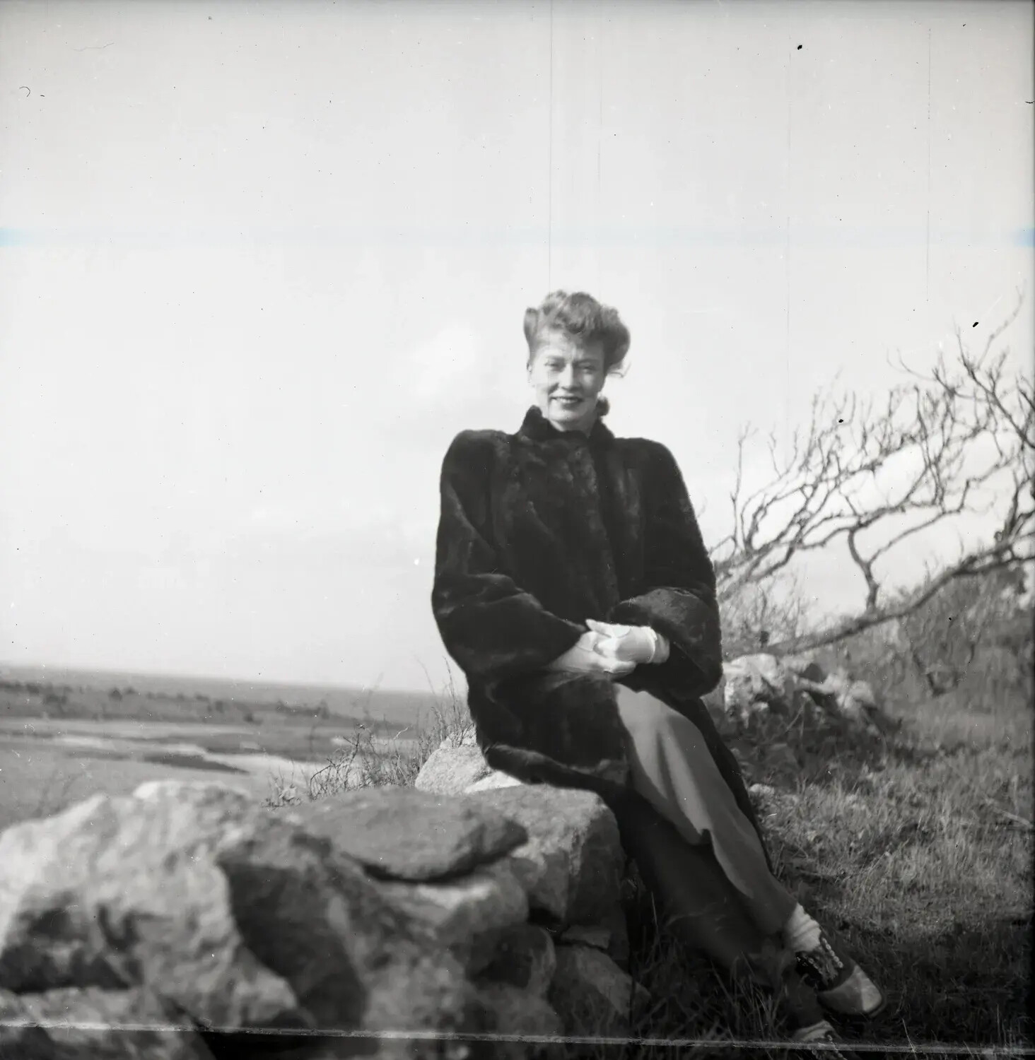 Woman in Fur Coat Seated on a Stone Wall Overlooking a Salt Marsh