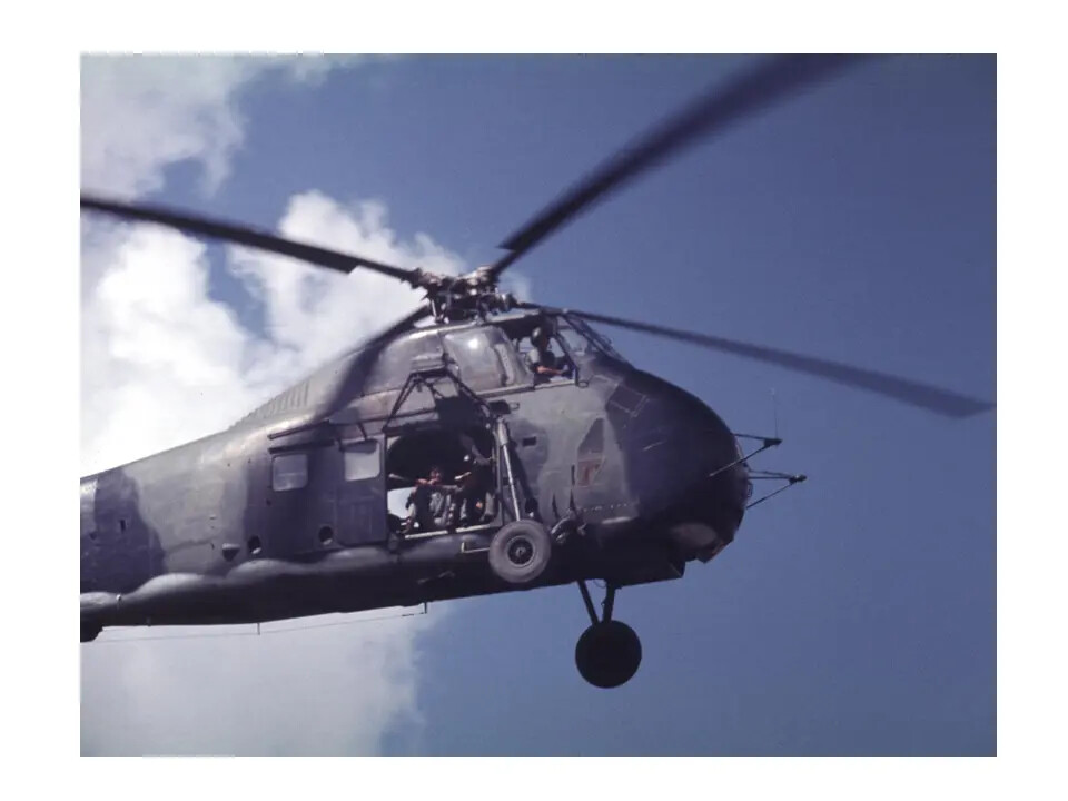 Camouflaged H-34 Helicopter in Flight, Crew Visible in Open Cargo Door