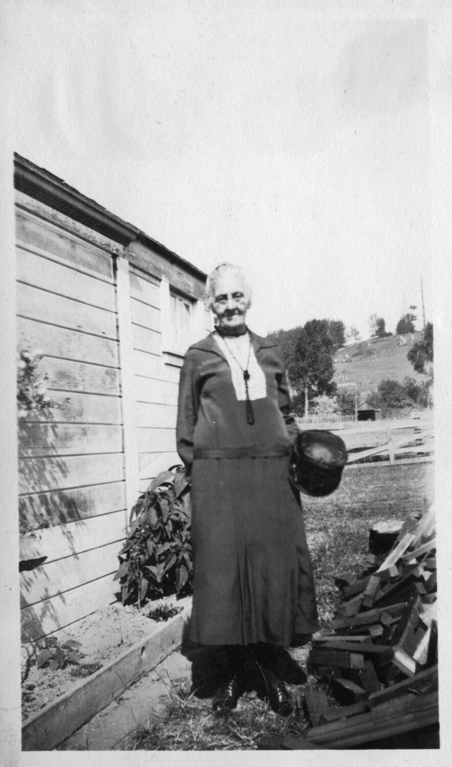 Elderly Woman in Dark Dress Beside Farmhouse and Woodpile, c. 1920s