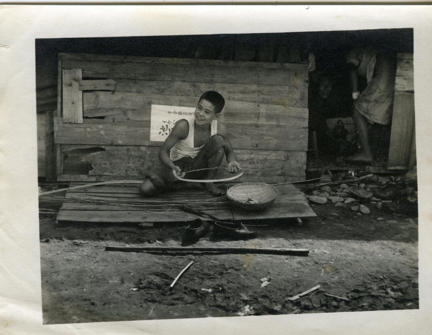 Okinawan Boy Weaving Bamboo Basketry Outside a Wooden Dwelling, Okinawa, 1945