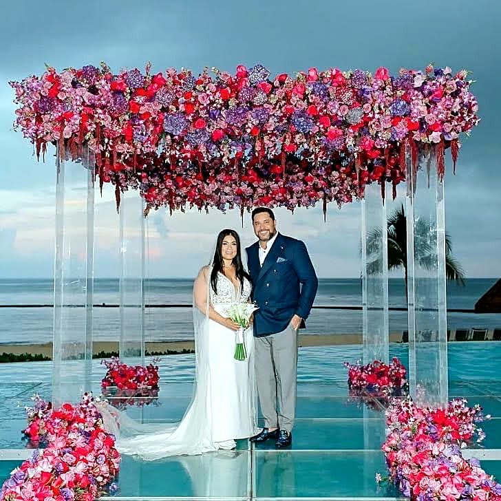 A wedding couple beneath colorful flowers on a platform overlooking the water.