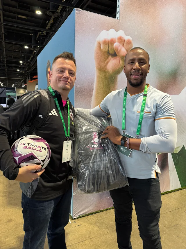 Two attendees displaying a soccer ball and Ultimateballa coaching bag at an indoor event