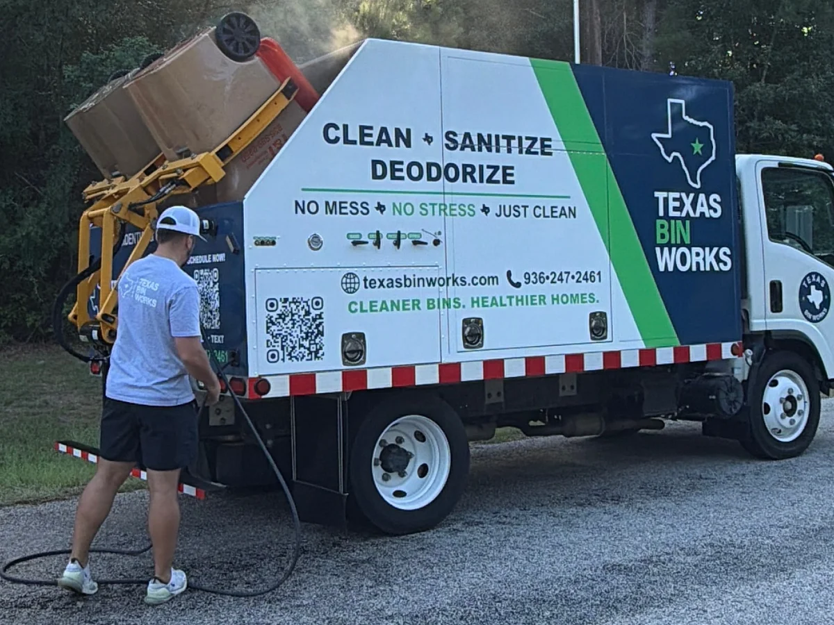 A Texas Bin Works truck with a bin cleaning mechanism actively steaming a trash can on a rural road.