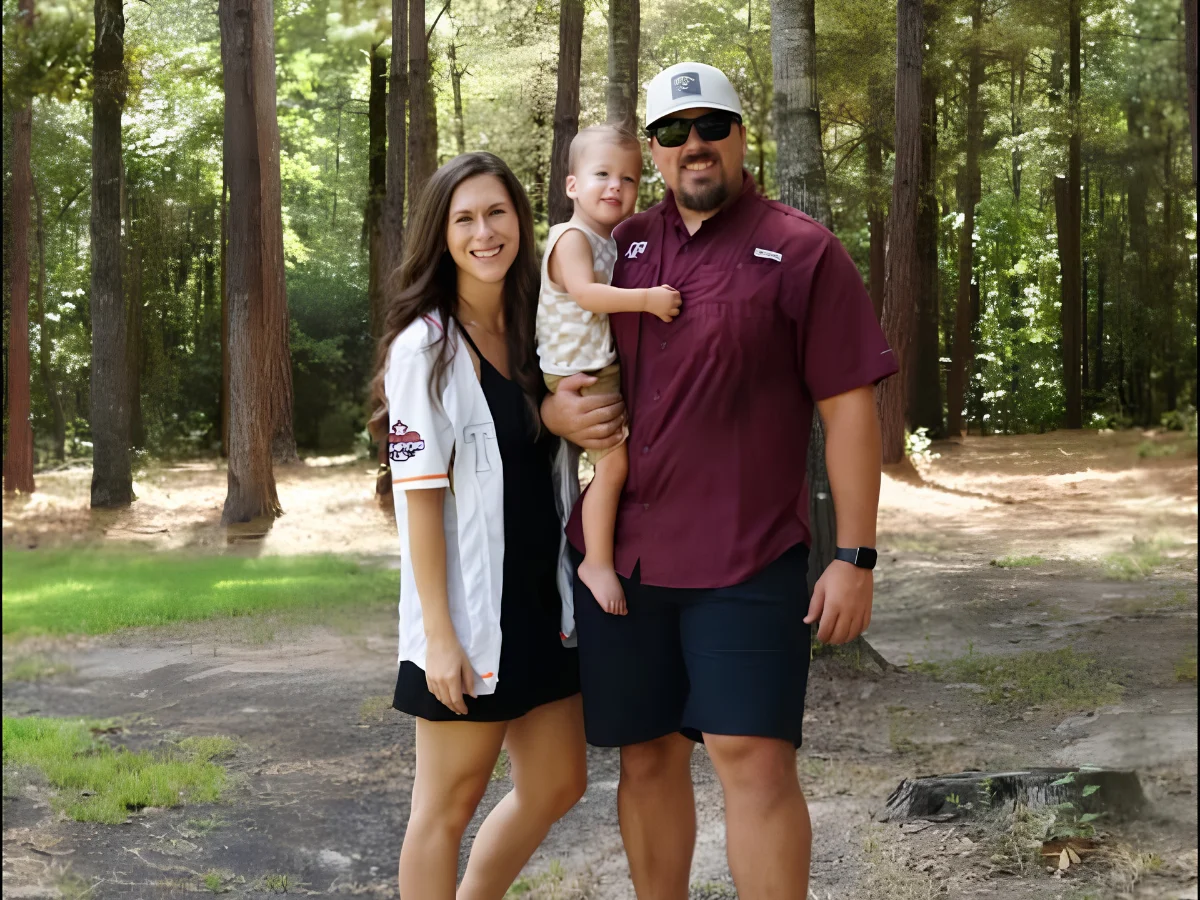 A smiling family of three, parents and their young child, pose outdoors in a sunlit wooded park.