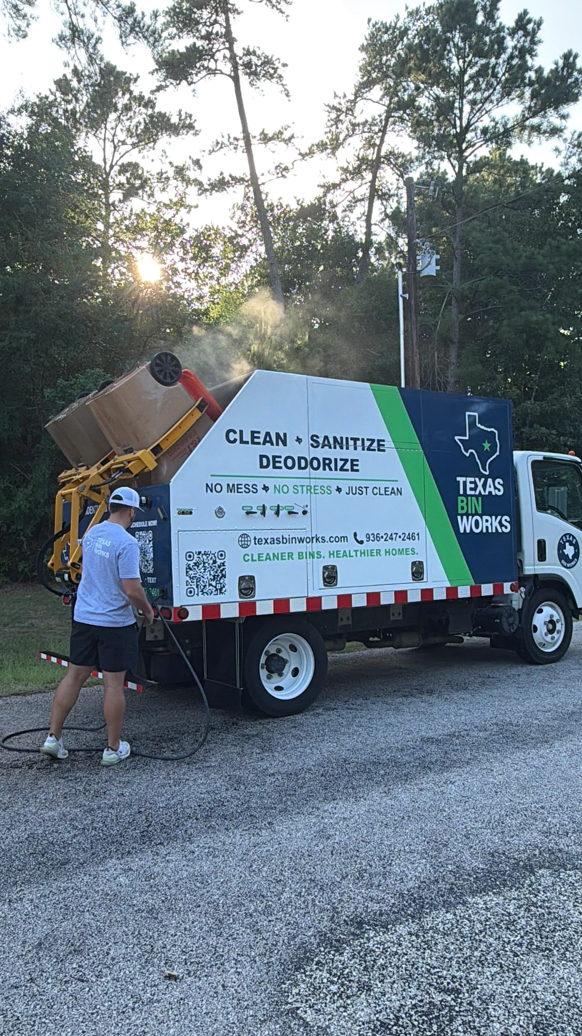 A Texas Bin Works truck cleaning a large trash bin with steam, operated by a person holding a hose.