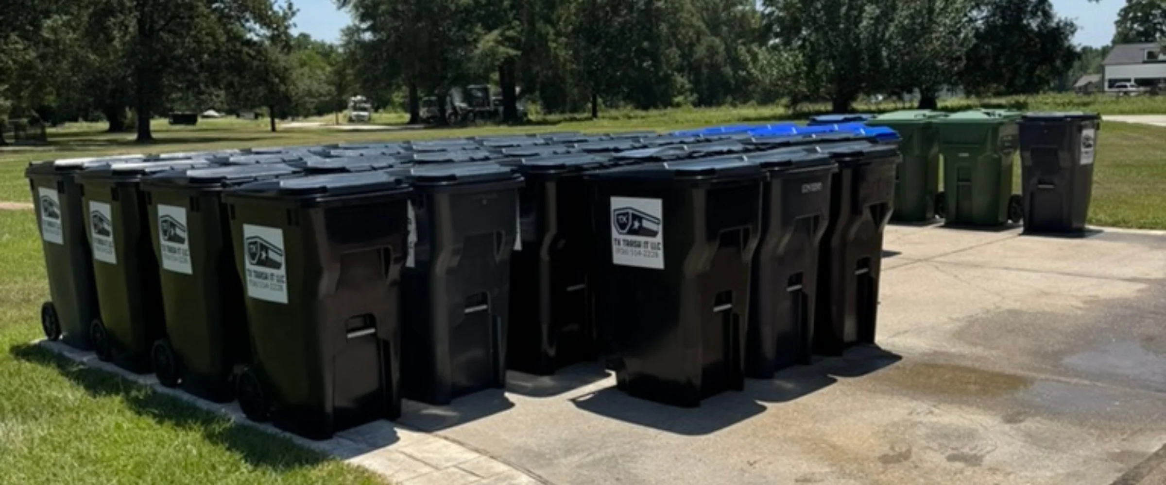 A long row of black and a few green garbage cans with 'TN TRASH IT LLC' logos, lined on concrete next to grass.
