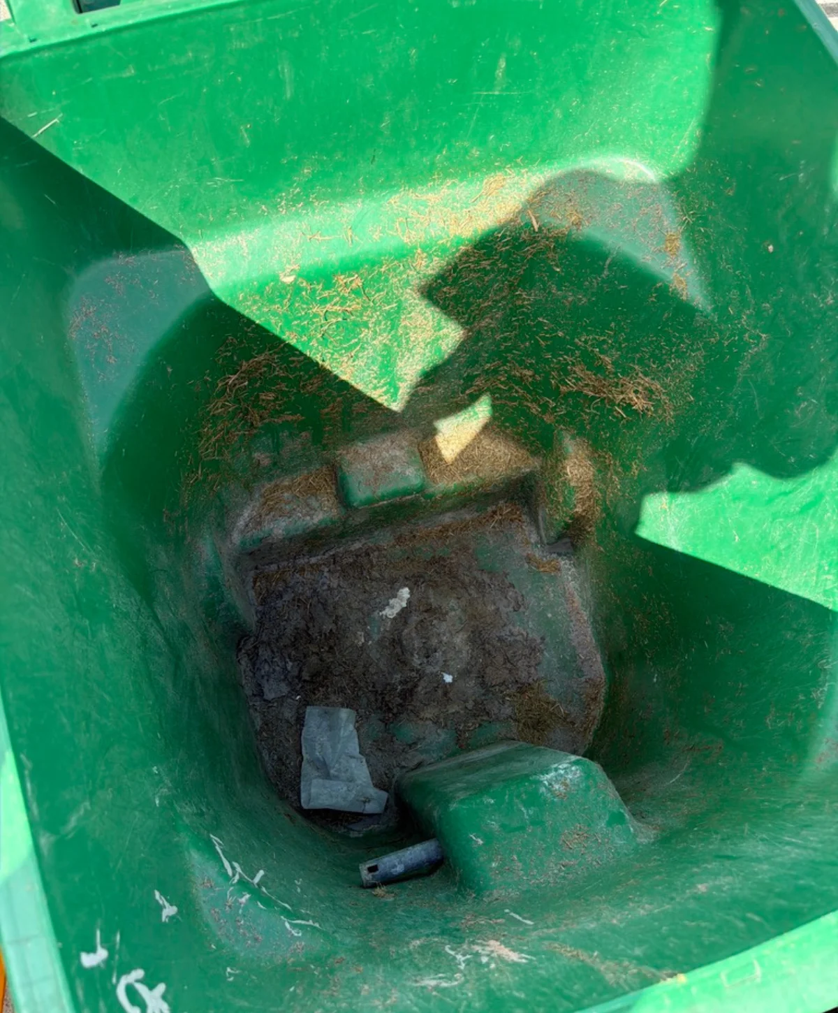 An overhead view looking inside a dirty, green trash bin with mud, grass clippings, and debris at the bottom.